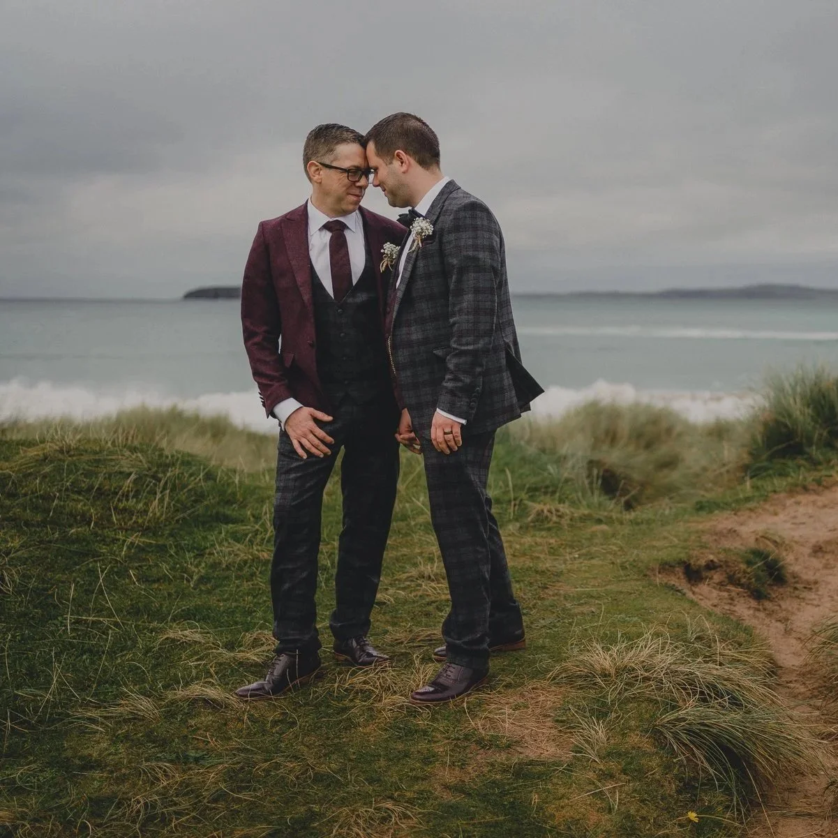 Two men in suits standing close together on a grassy area by the beach, with overcast sky and ocean in the background, sharing an intimate moment.