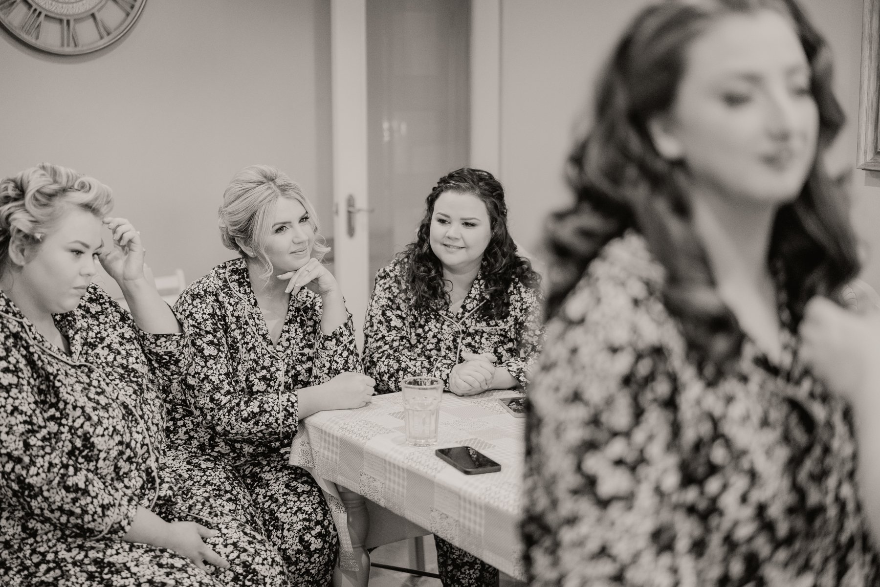 Four women sitting at a table, dressed in matching floral pajamas, with a glass of water and a phone on the table, preparing for a wedding or event, with one woman in the foreground looking at her reflection.