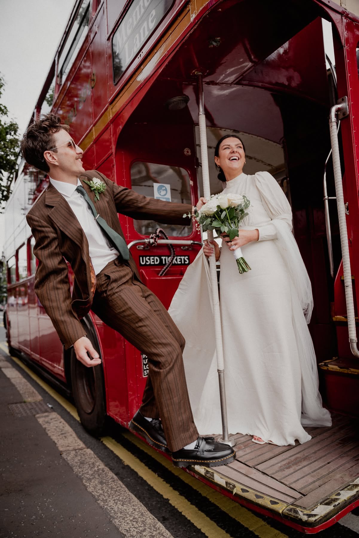 A man in a brown striped suit and a woman in a white wedding dress are standing on a red double-decker bus, smiling and holding flowers. The man is wearing sunglasses and leaning towards the woman, who is holding a bouquet of white roses. They appear to be celebrating or taking a photo on a city street.