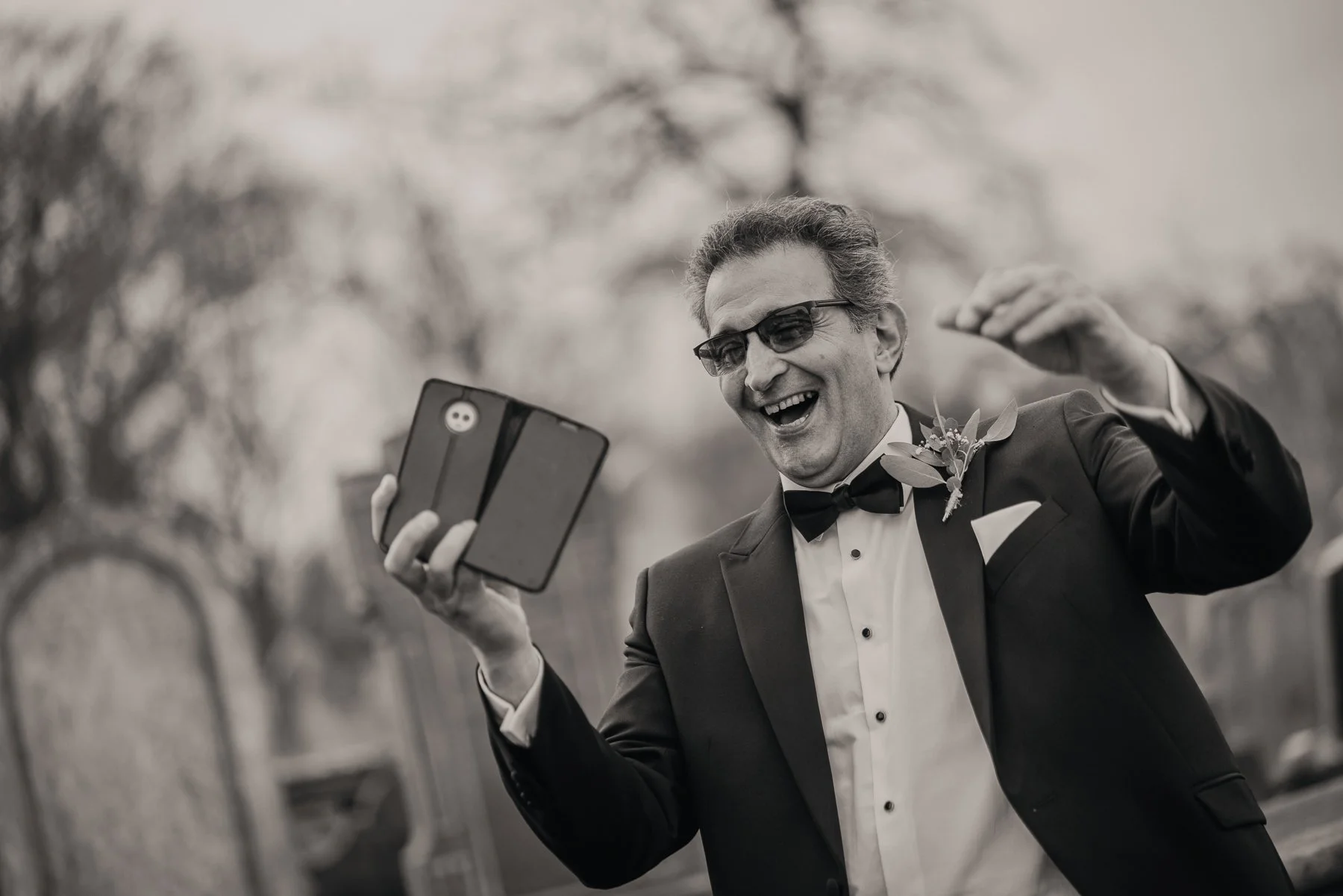A man in a tuxedo with a bow tie and glasses, smiling joyfully as he takes a selfie outdoors.