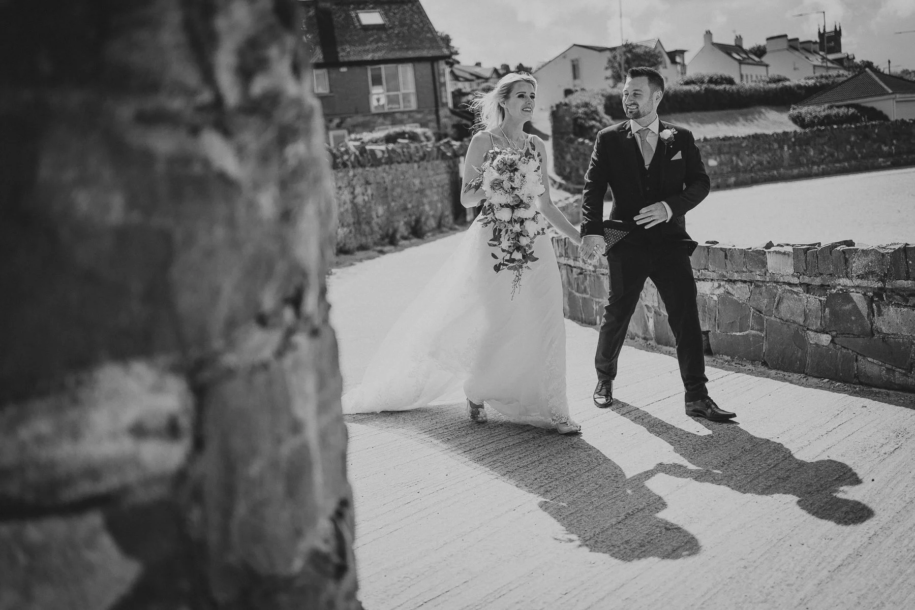 A black and white photo of a bride and groom walking hand in hand along a sidewalk near a river, with houses in the background. The bride is wearing a wedding dress and holding a large bouquet, and the groom is dressed in a suit with a boutonniere. T