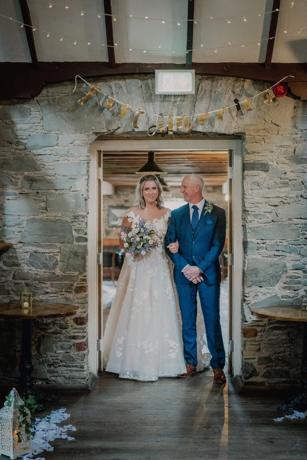 A bride in a white wedding gown holding a bouquet and a groom in a blue suit standing in a rustic stone doorway decorated with a "Just Married" banner, inside a cozy venue.