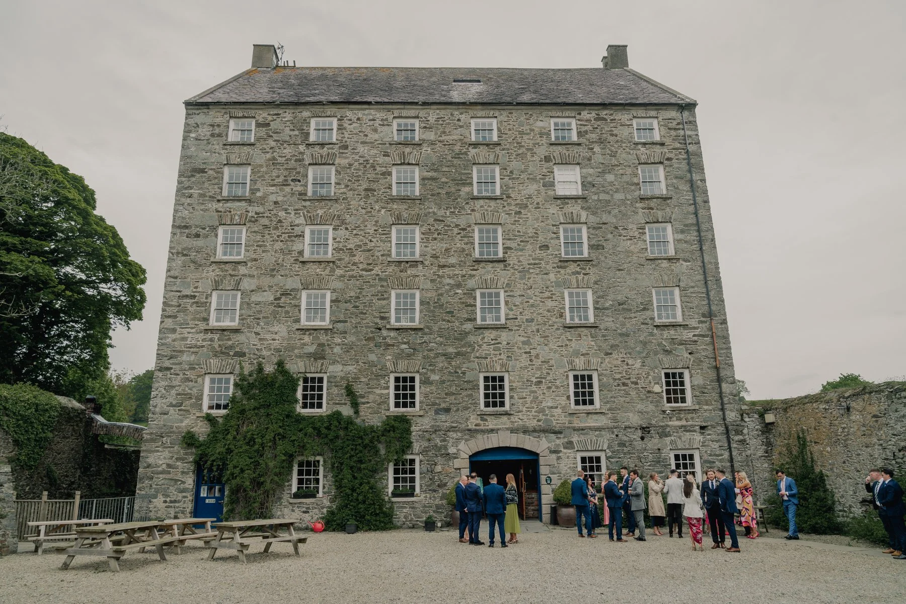 A large stone historic building with multiple windows and a few chimneys on the roof. There is a group of people dressed in formal attire gathered outside, some talking and others waiting. The setting appears to be an outdoor event at the building, w