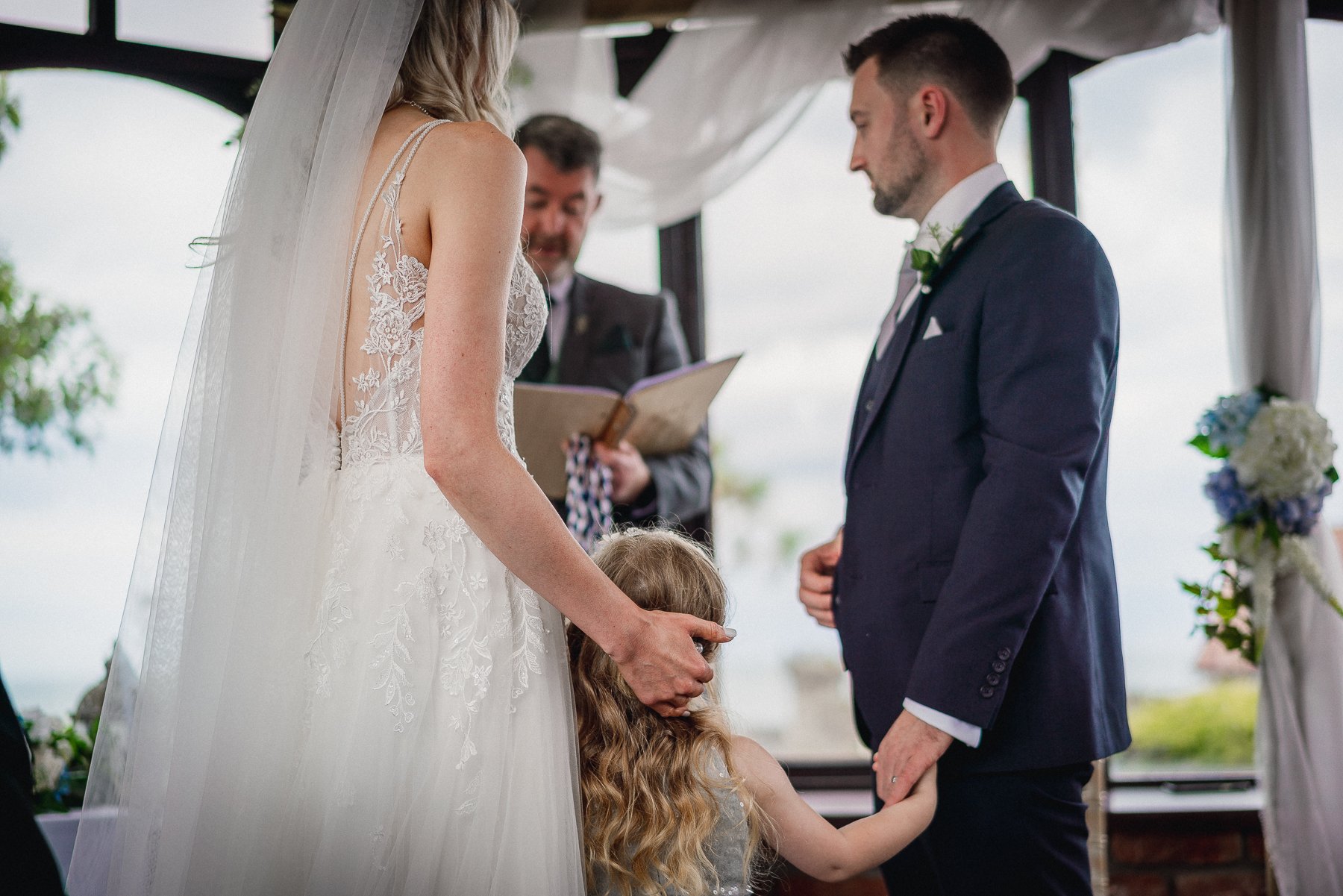 A couple getting married outdoors, with a woman in a lace wedding dress and a man in a navy suit, are holding hands and facing each other while a officiant officiates the ceremony. A young girl with long hair is holding the woman's hand. The ceremony