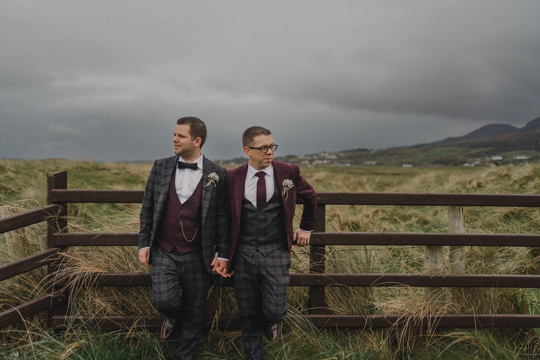 Two men dressed in plaid suits and ties, holding hands, standing beside a wooden fence in a grassy outdoor area under a cloudy sky.