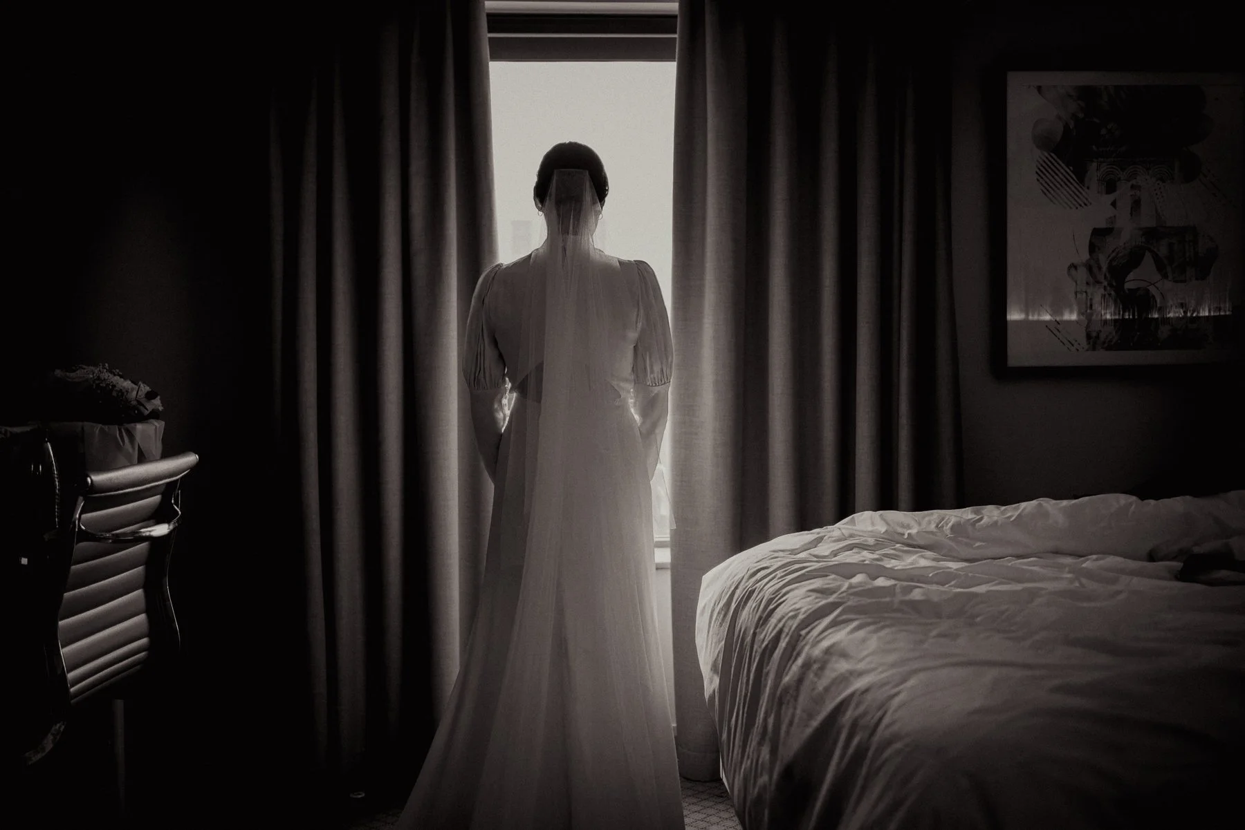 A woman in a long wedding dress and veil stands in front of a window with curtains. The room appears dark, with a bed on the right and a nightstand on the left with flowers. The scene has a contemplative or bridal mood.