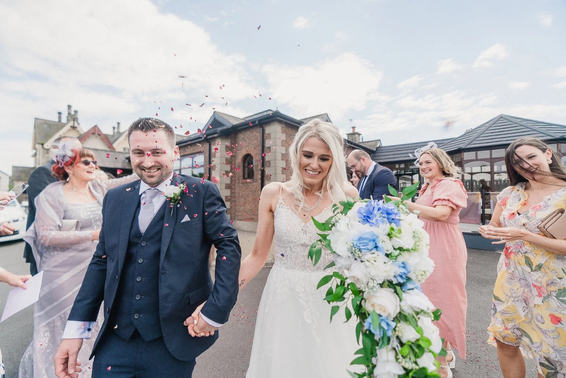 Bride and groom holding hands and walking after their wedding, surrounded by smiling friends and guests, with confetti falling in the air, outdoors with a building and cloudy sky in the background.