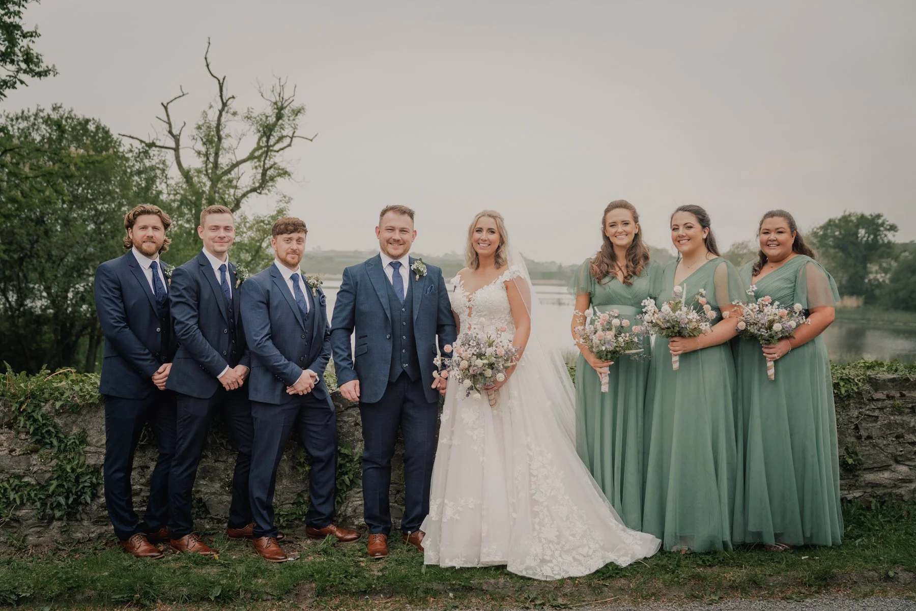 A group of eight people dressed in wedding attire standing outdoors near a body of water, with trees in the background. The bride and groom are in the center, surrounded by four bridesmaids in green dresses and two groomsmen in suits. Everyone is smi