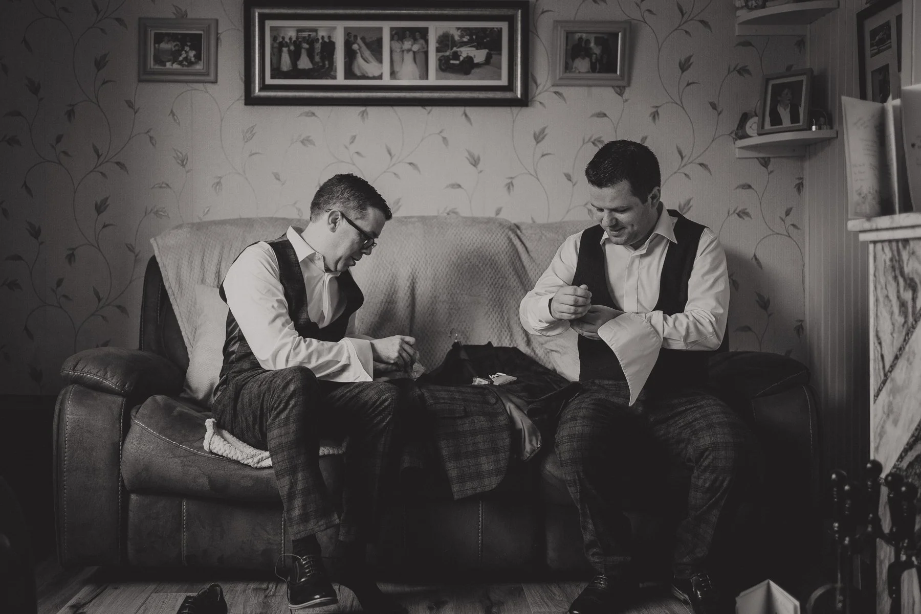Two men in formal attire sitting on a couch in a living room, looking at their phones and smiling. The room has framed photos on the wall, including a large wedding photo collage, and decorative wallpaper with a leaf pattern.
