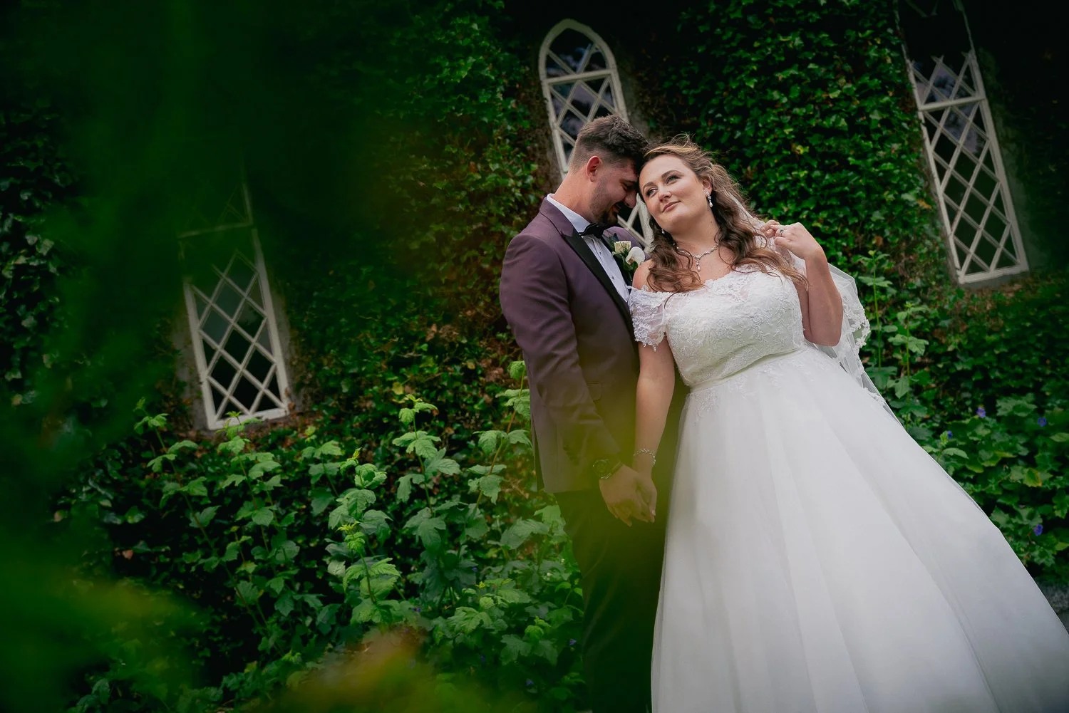 A bride and groom holding hands and showing affection on their wedding day outdoors, with green foliage and windows in the background.