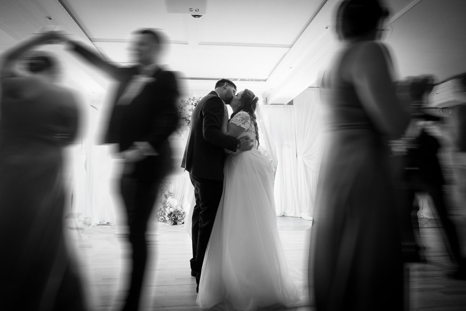 A black and white photo of a newlywed couple kissing on the dance floor, with blurred guests dancing around them.