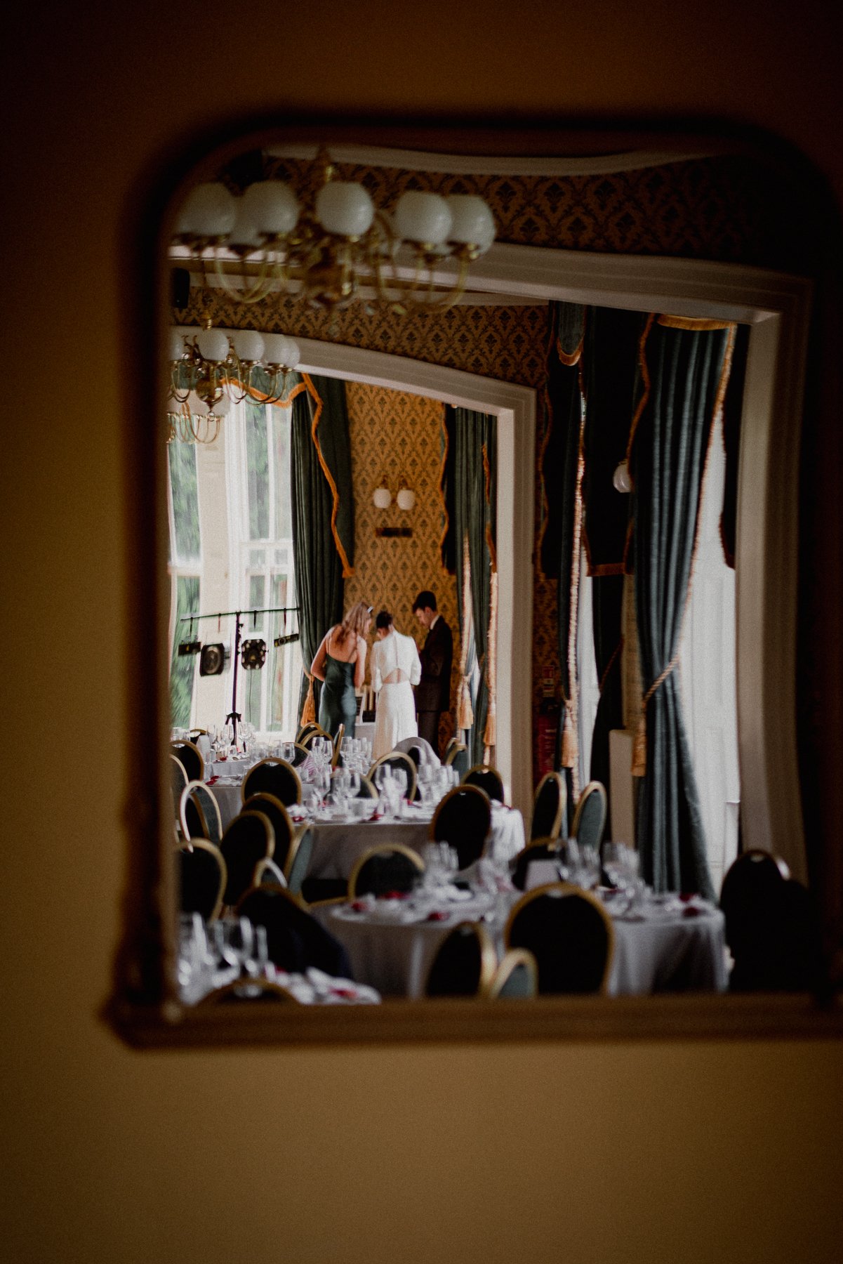 A view through a gap in a wall shows an elegantly decorated dining room with round tables set for an event. Three people, a woman in a dark dress, a woman in a white dress, and a man in a suit, are gathered and talking near a window with heavy, dark 