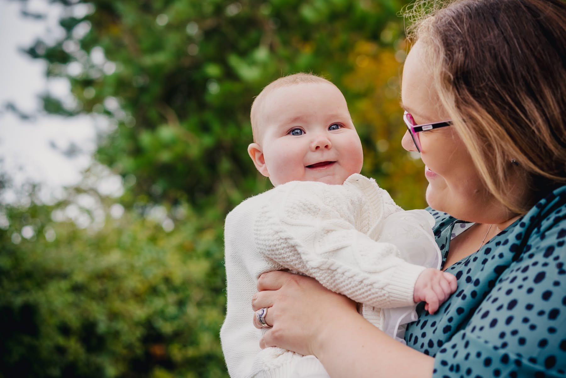 A woman with brown hair and glasses holding a smiling baby girl outdoors with green trees in the background.