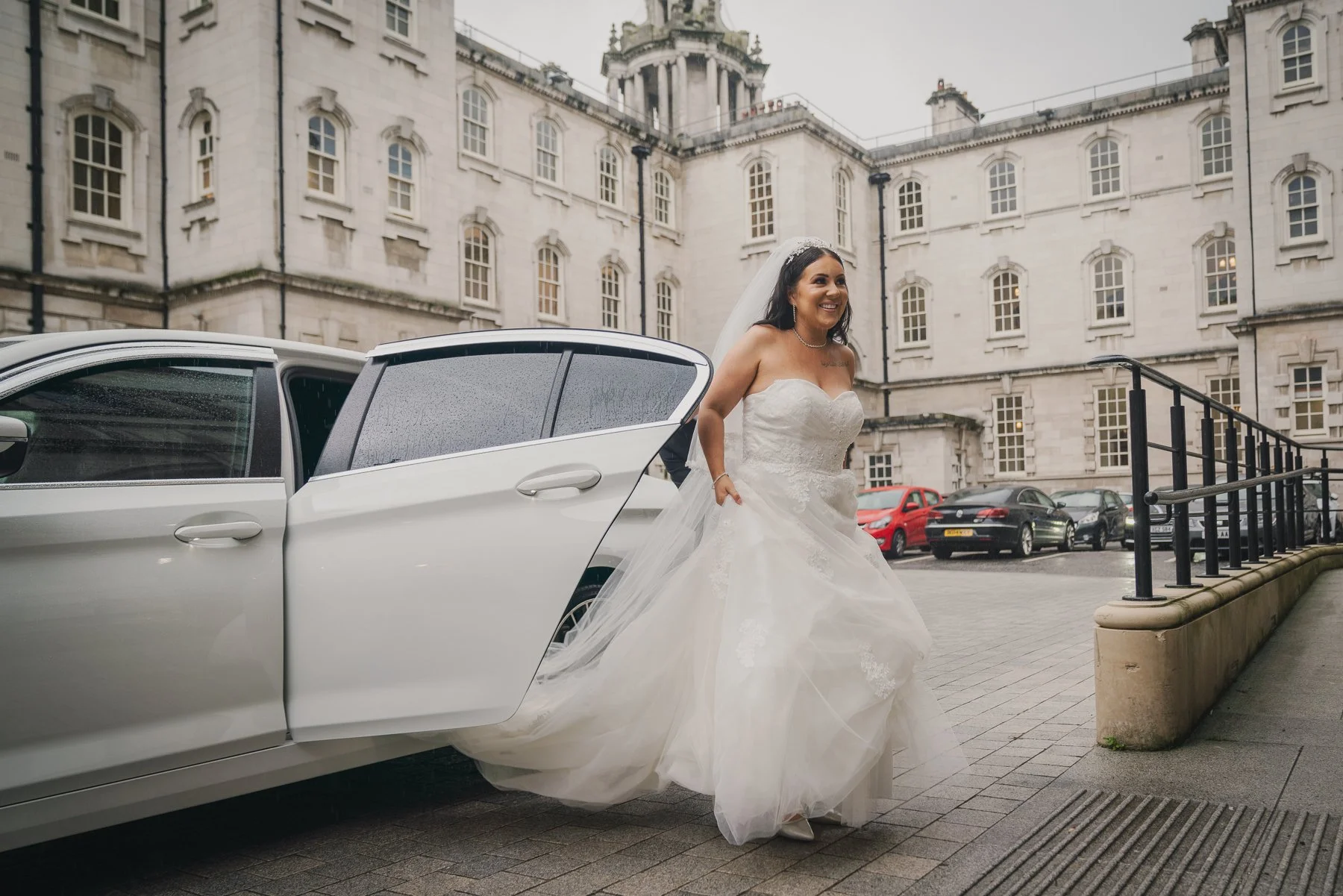 A bride in a white wedding dress arriving at a building, exiting a white limousine with the door open, on a paved area beside parked cars, with an ornate building in the background.