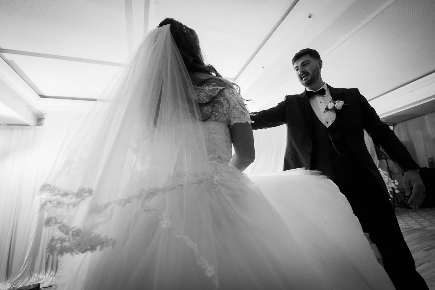 Black and white photo of a groom in a tuxedo reaching out to a bride in a wedding gown with a veil, standing next to a large wedding cake, in a decorated indoor setting.
