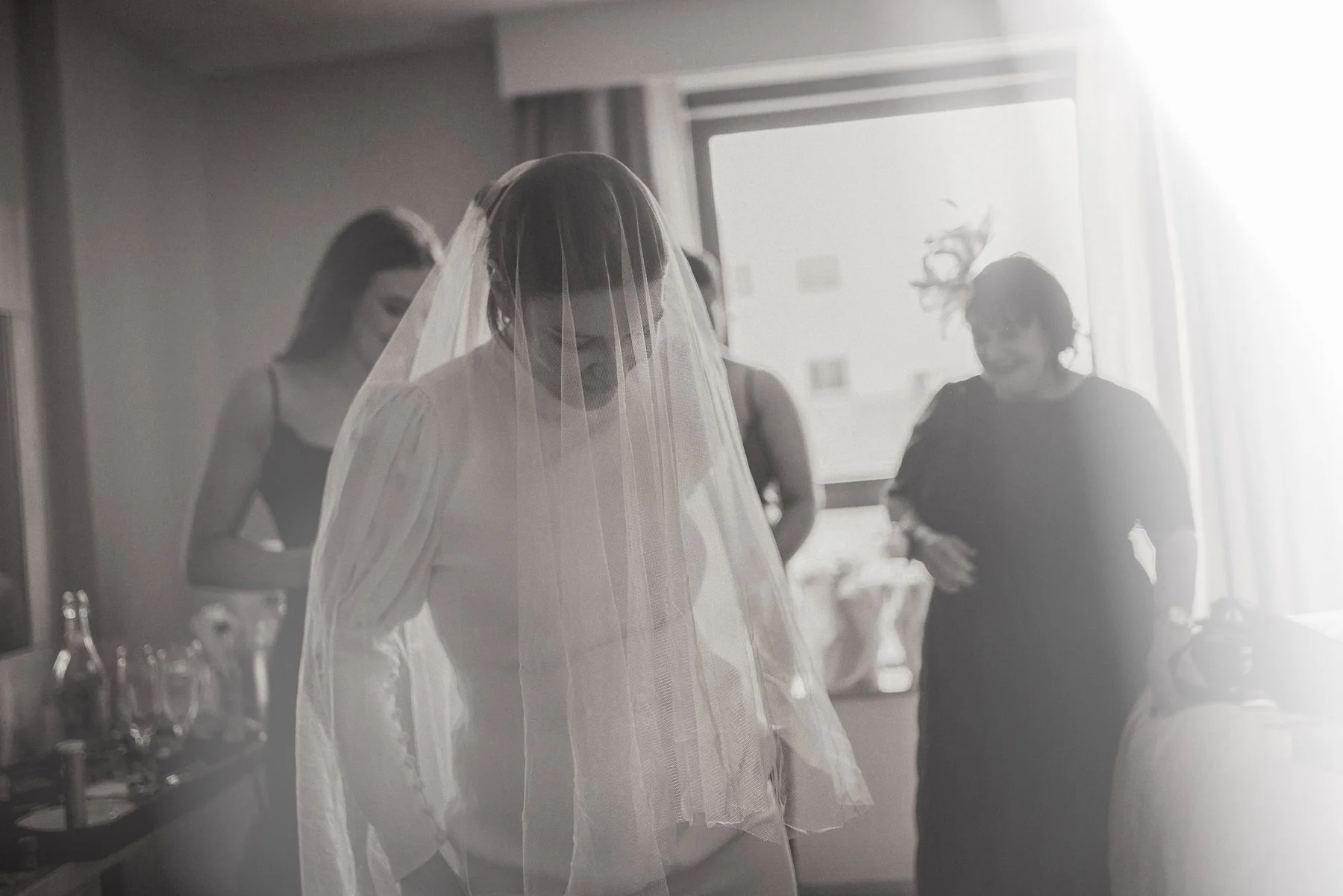 A bride wearing a veil and wedding dress standing with her head bowed, surrounded by women in black dresses in a softly lit room.
