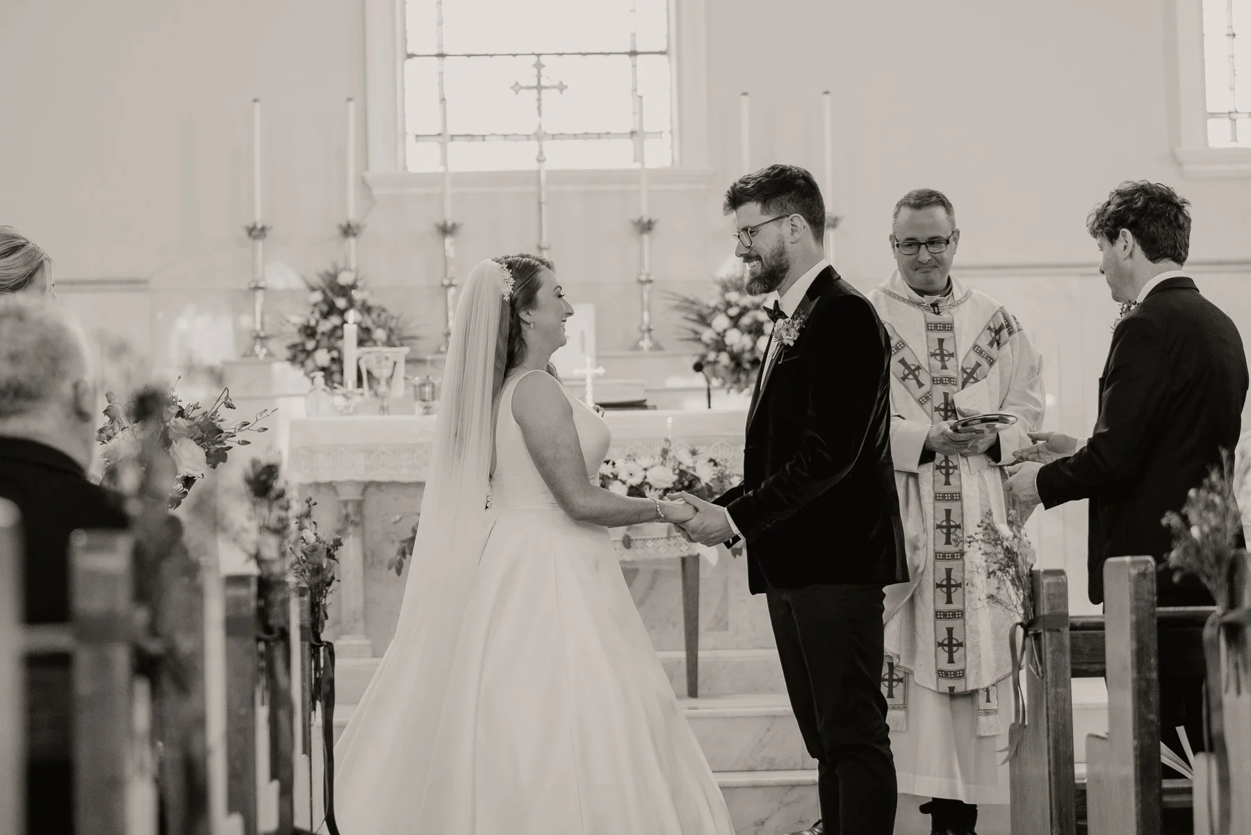 A bride and groom exchange vows during a wedding ceremony inside a church, holding hands and smiling at each other, with a priest officiating behind them.