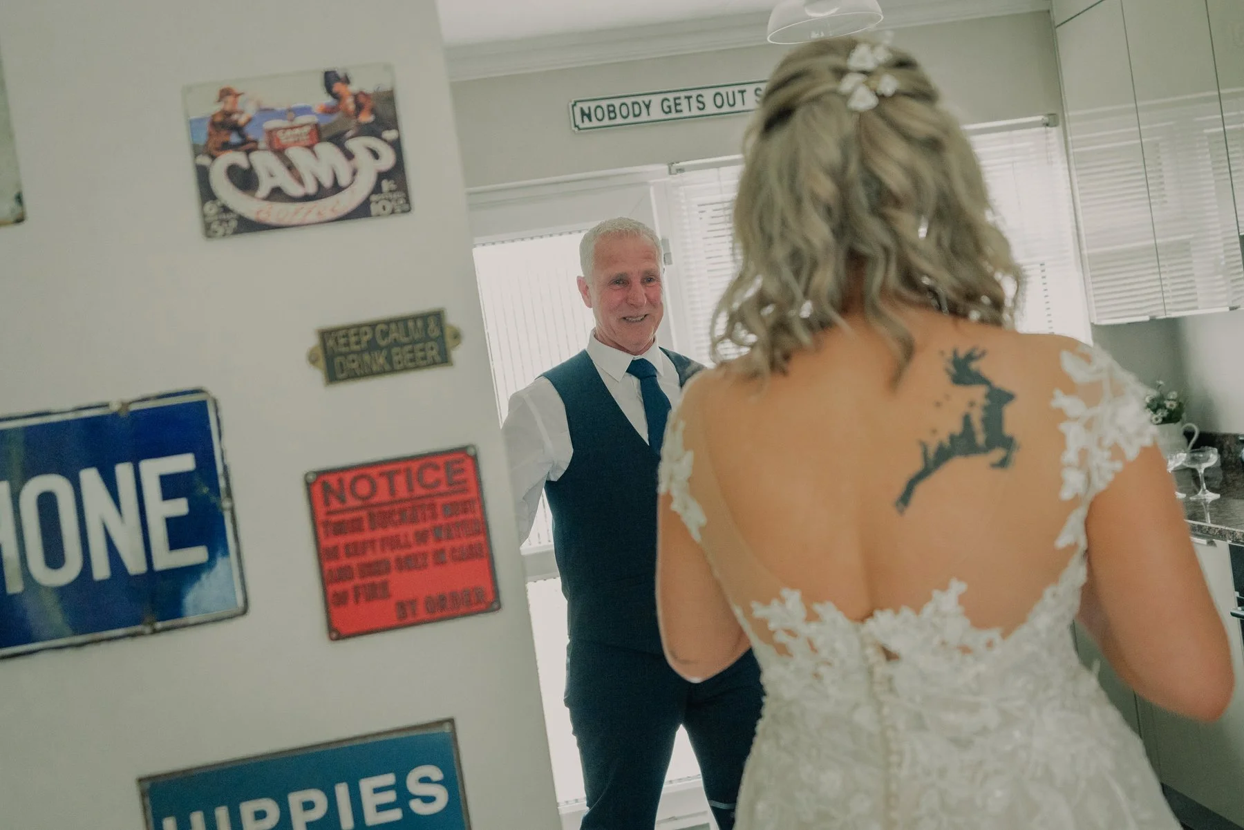 A woman in a white lace wedding dress with a black deer silhouette tattoo on her upper back is standing in front of an older man in a vest and tie, smiling. The image is taken in a room with signs on the wall, including one that says 'NOBODY GETS OUT