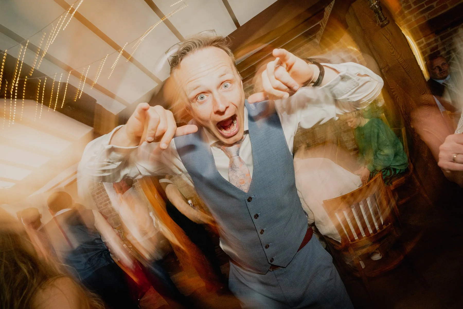 Excited man in a gray vest and patterned tie at a social gathering, pointing towards the camera with a lively expression, in a dimly lit restaurant or bar with wooden furniture.