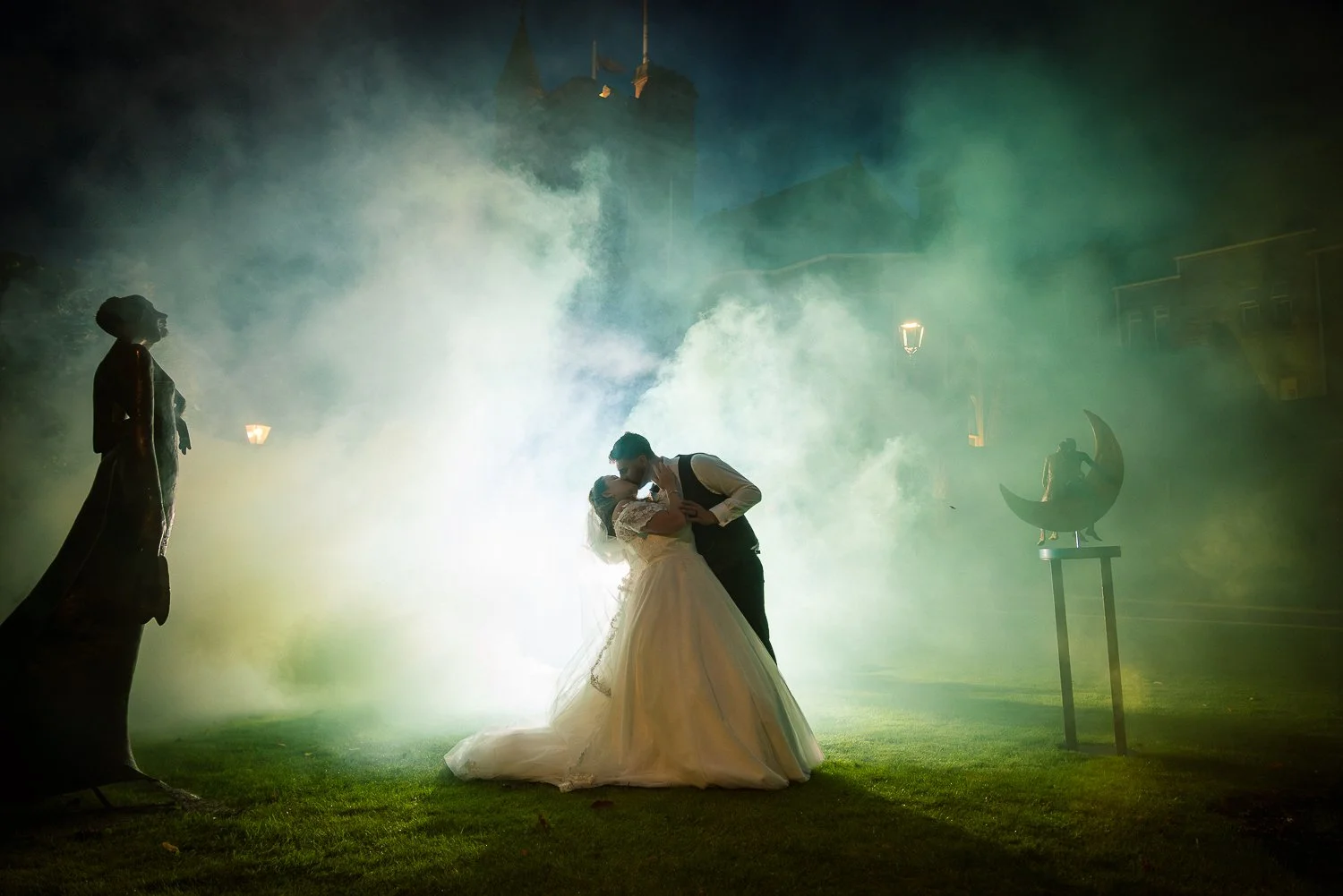 A bride and groom share a kiss during their wedding with fog and soft lighting, a statue of a witch on a broomstick, and a castle in the background.