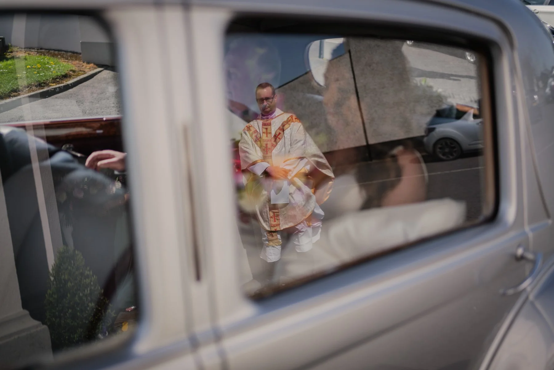A priest dressed in ornate vestments is reflected in the side mirror of a vehicle, standing outdoors near a parking lot.