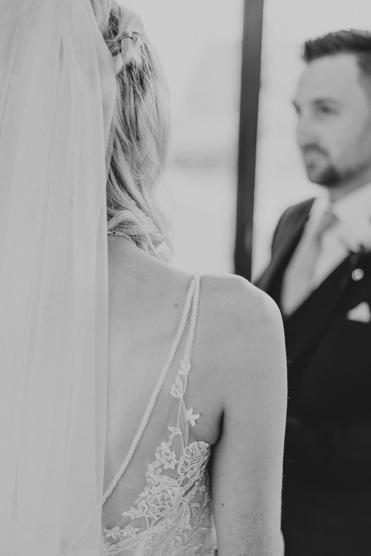 A bride wearing a lace wedding dress with spaghetti straps, her back partly turned, with her hair styled in loose curls. An officiant, dressed formally, stands behind her and out of focus.