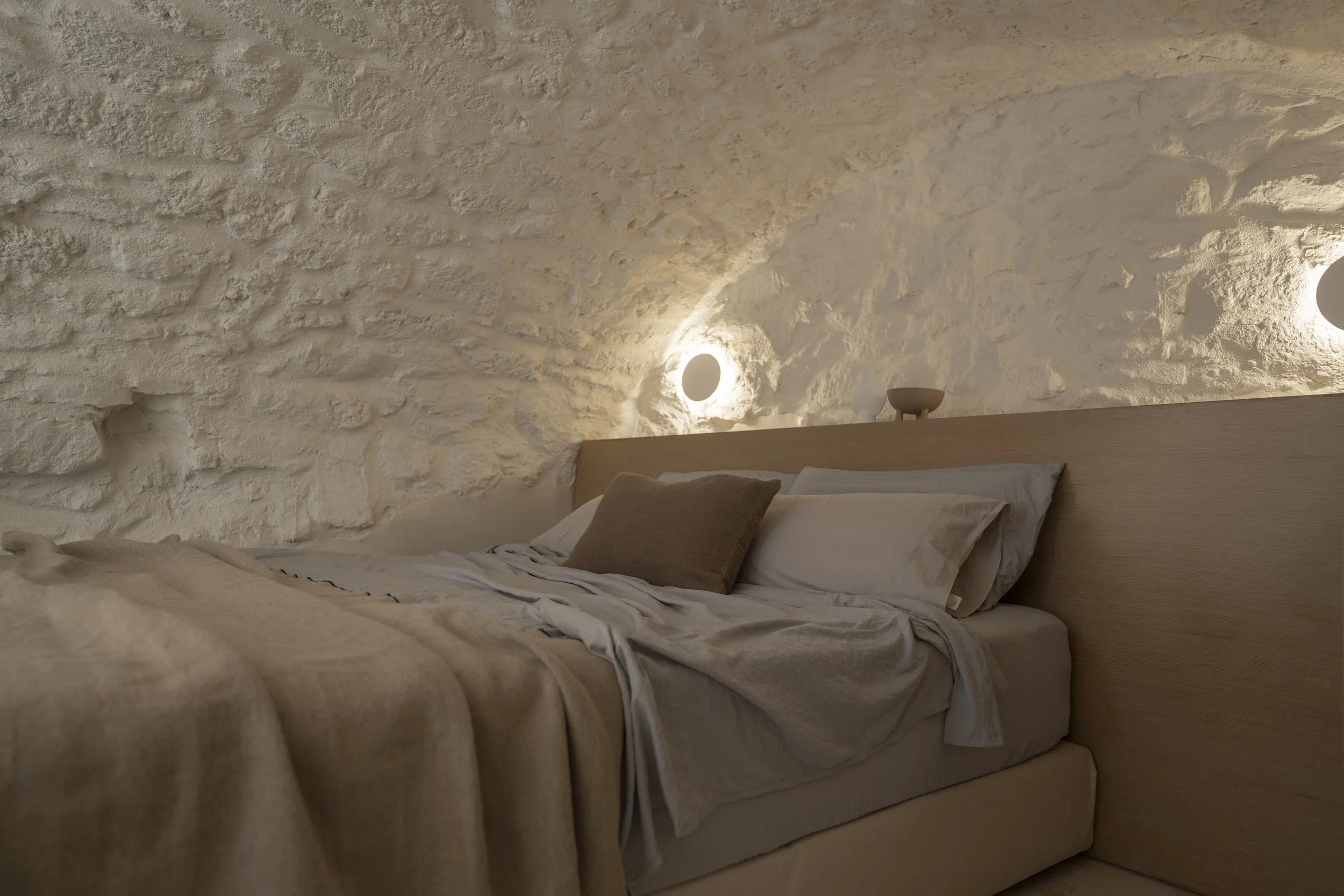 A cozy bedroom with a textured white stone wall, a bed with white and beige bedding, and warm wall-mounted lights.