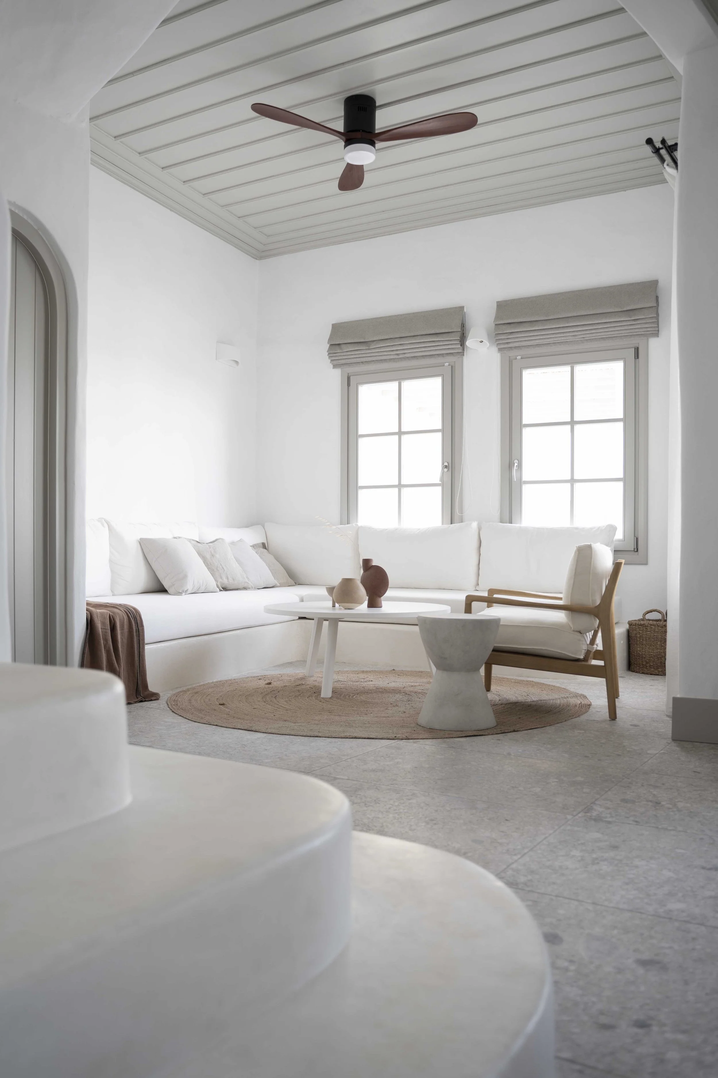 A bright and minimalist living room with a white sofa, beige pillows, and wooden accents. There are two windows with beige window shades, a round woven rug, and a modern coffee table with decorative vases. The ceiling has a wooden fan and the walls are white, creating a clean and airy atmosphere.