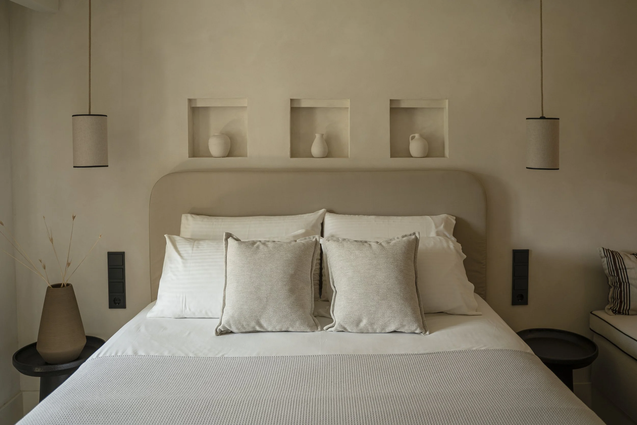 A neatly made bed with white linens and multiple beige pillows, flanked by black bedside tables with spherical lamps and vases in built-in wall shelves above the headboard.