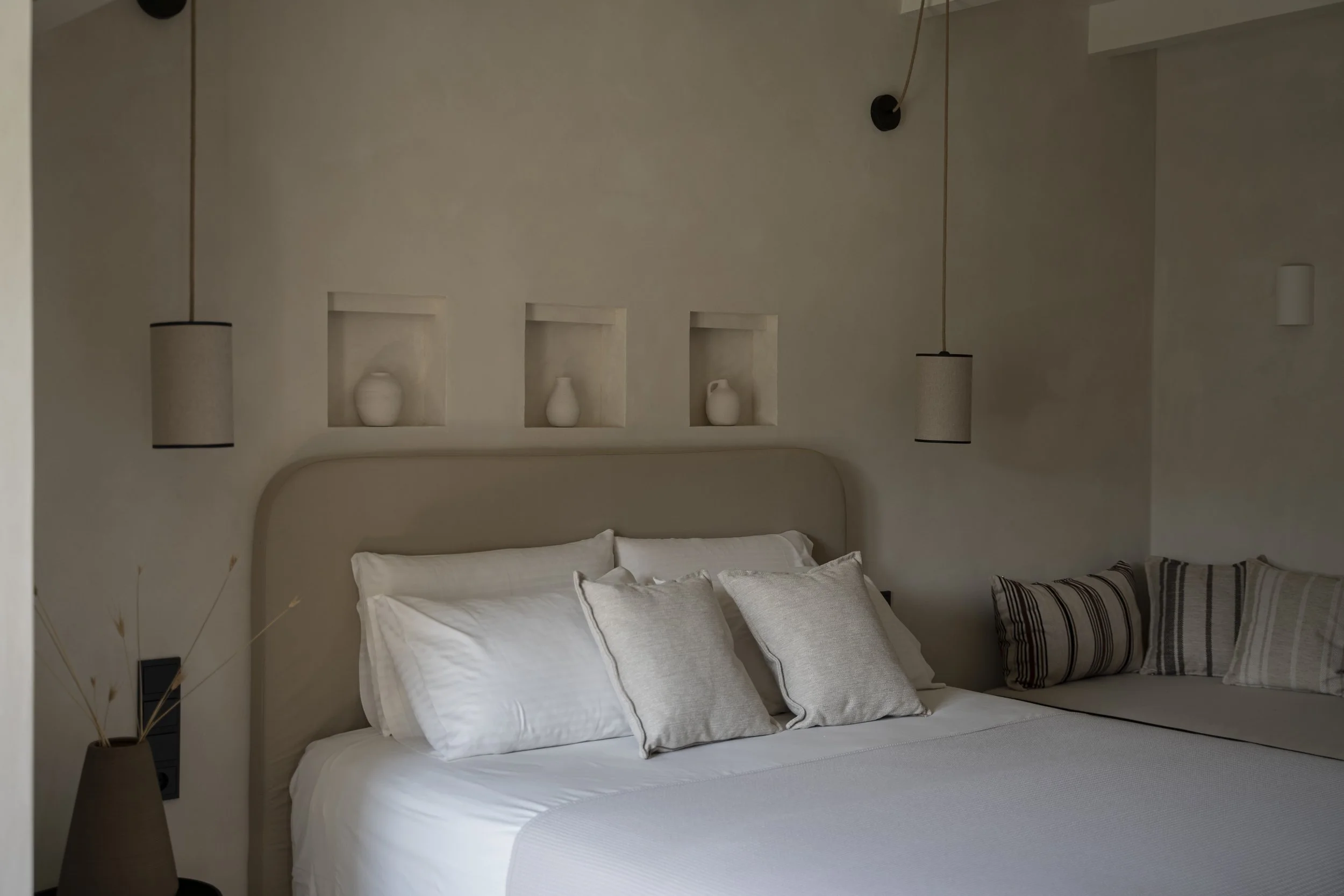 A neatly made bed with white bedding and several pillows in front, decorated with a black and white striped pillow. Behind the bed, the wall features three recessed niches, each containing a white decorative vase, and two hanging pendant lights with beige shades are on either side of the bed. A small vase with dried plants is visible on the bedside table on the left.