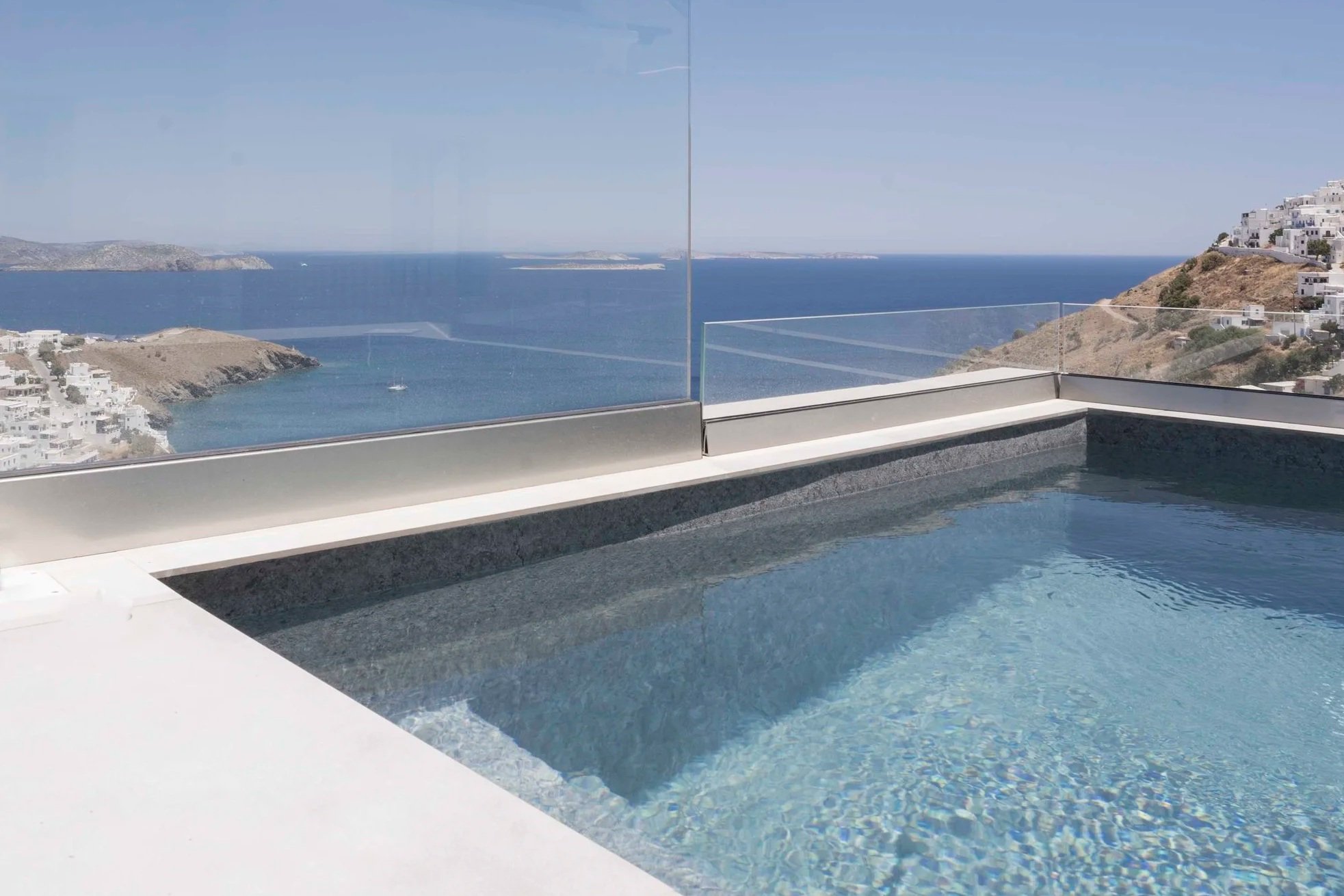 View from a balcony overlooking the ocean with hills and white buildings under a clear sky.