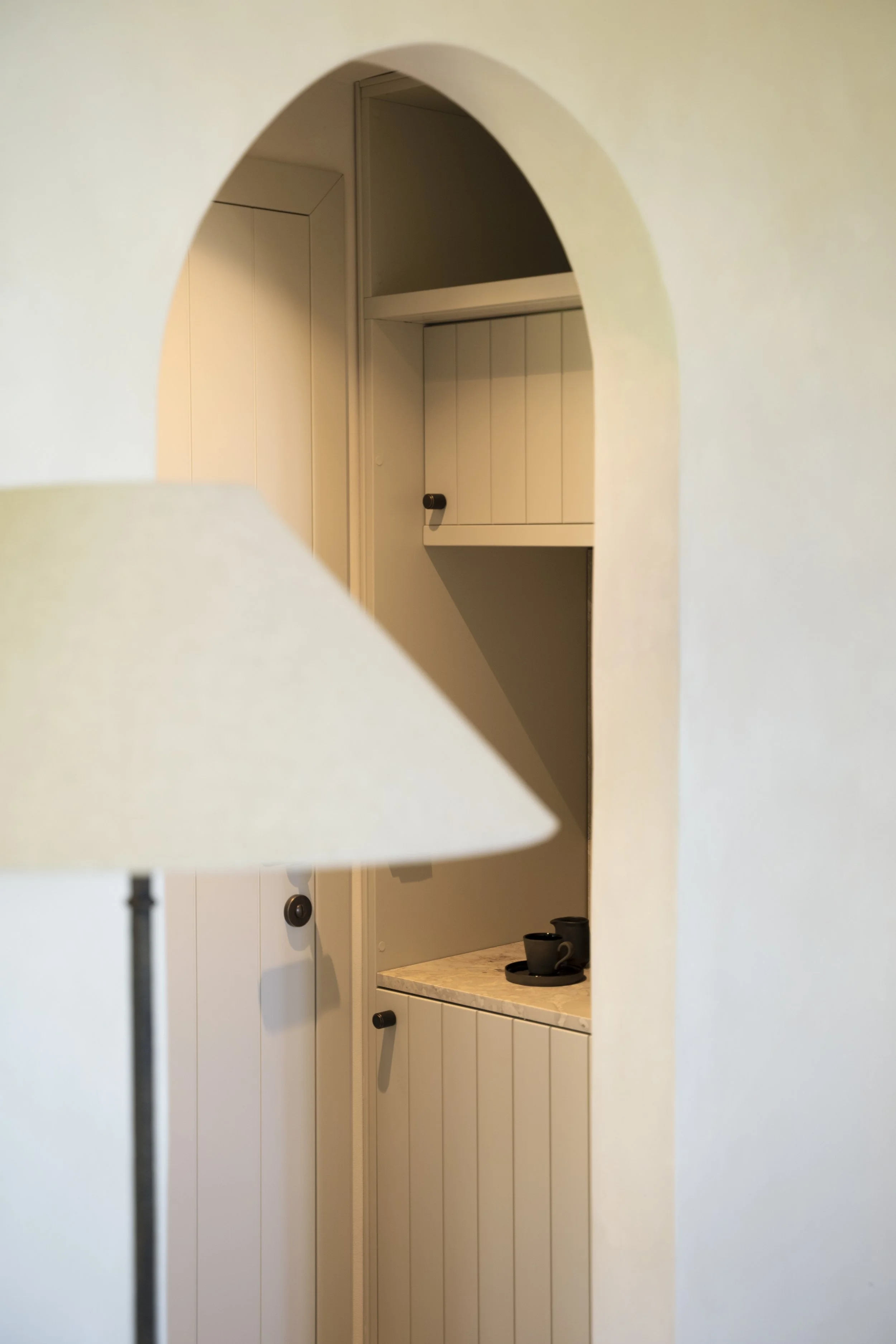 View of a small kitchen with white cabinets, a marble countertop, and black cups on a tray. The photo is taken through an arch-shaped opening in a white wall.