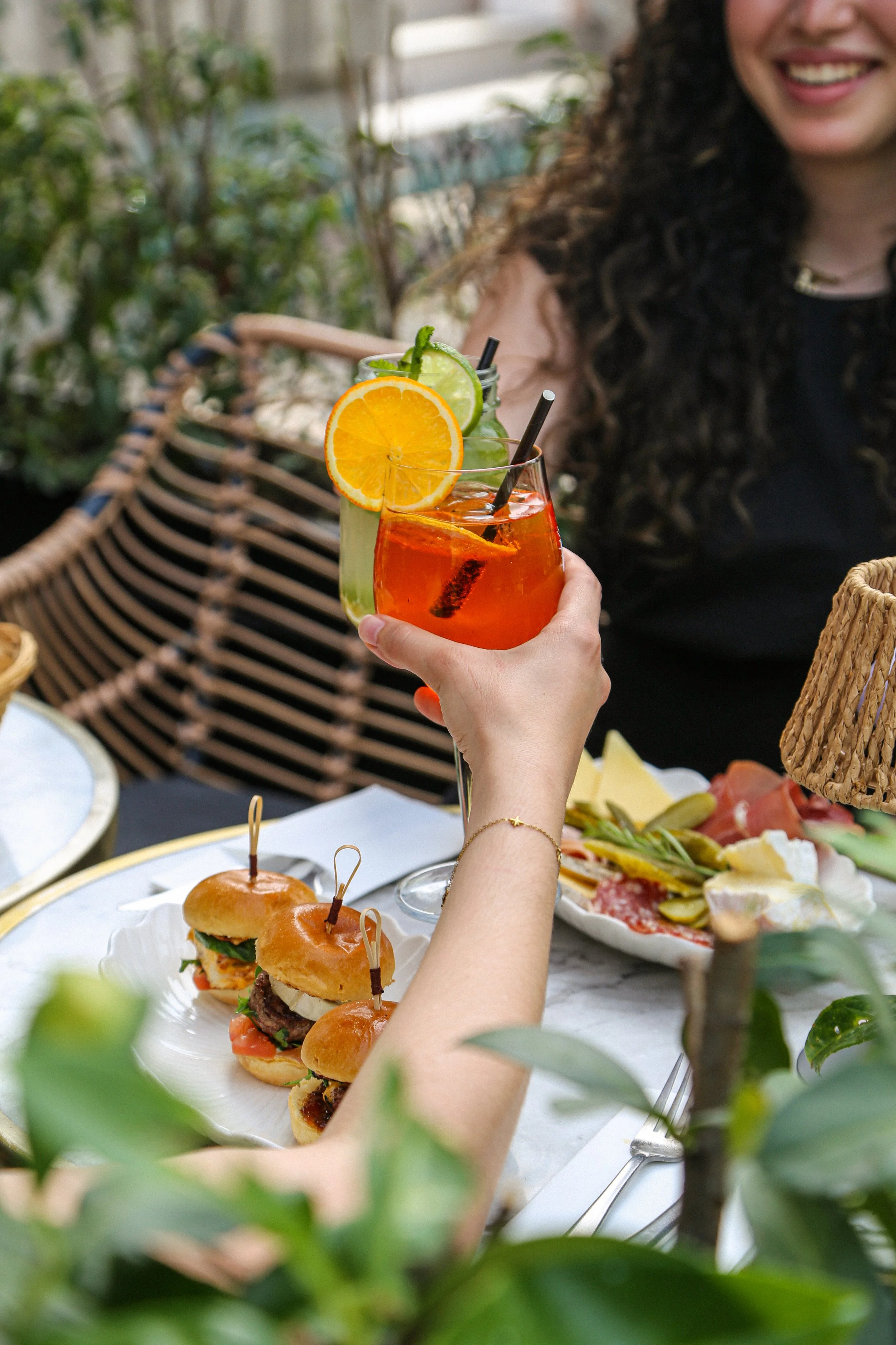 Verre de cocktail orange et citron avec une tranche d'orange et de citron, entouré de personnes où l'on voit partiellement une femme souriante avec de longs cheveux bouclés, à une table avec des amuse-bouches, un plateau de pizza et un sandwich, dans