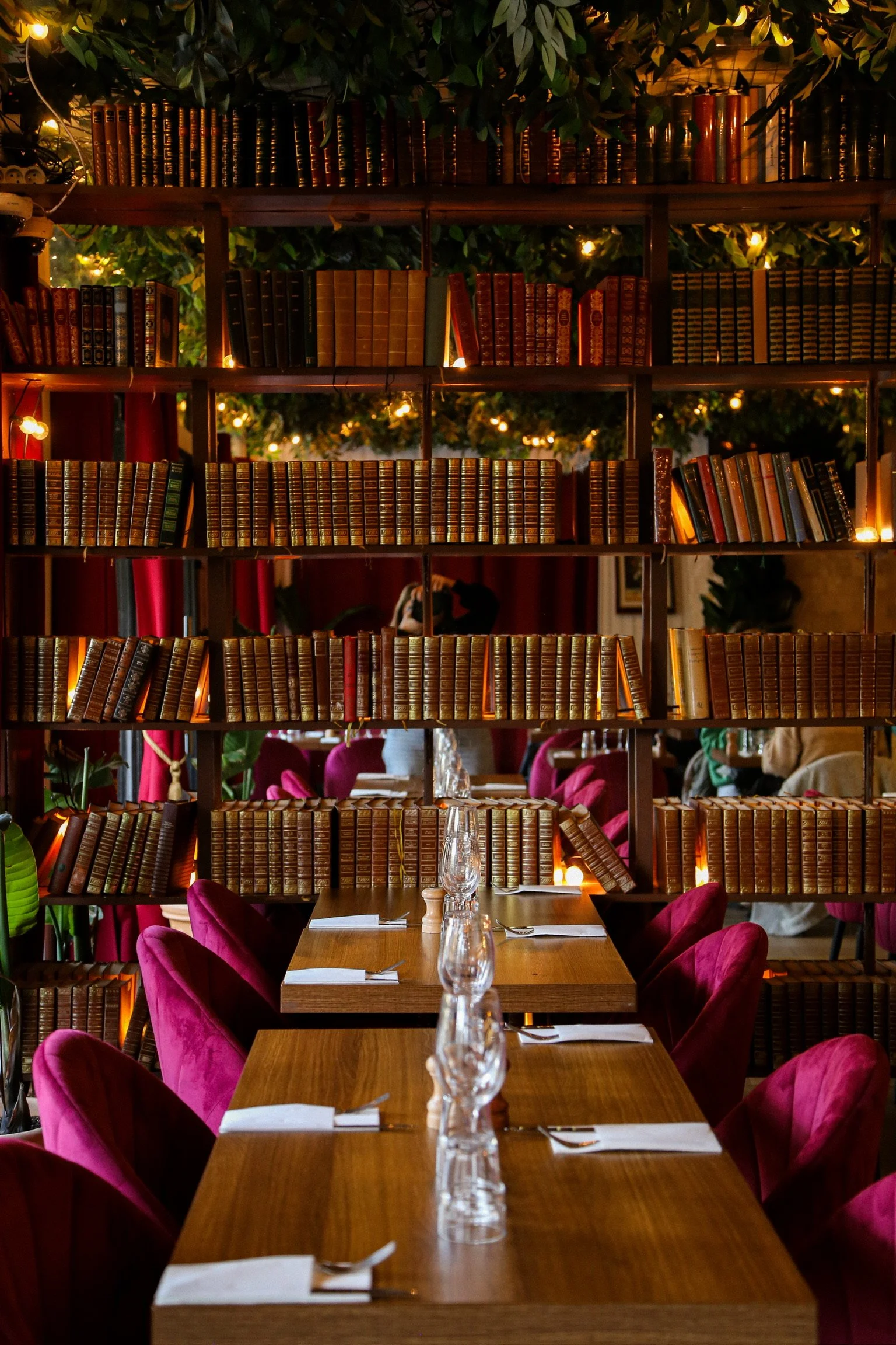 Intérieur d'un restaurant avec étagère en bois remplie de livres, lampes chaudes, tables avec glass et assiettes, chaises en velours baisé, ambiance chaleureuse
