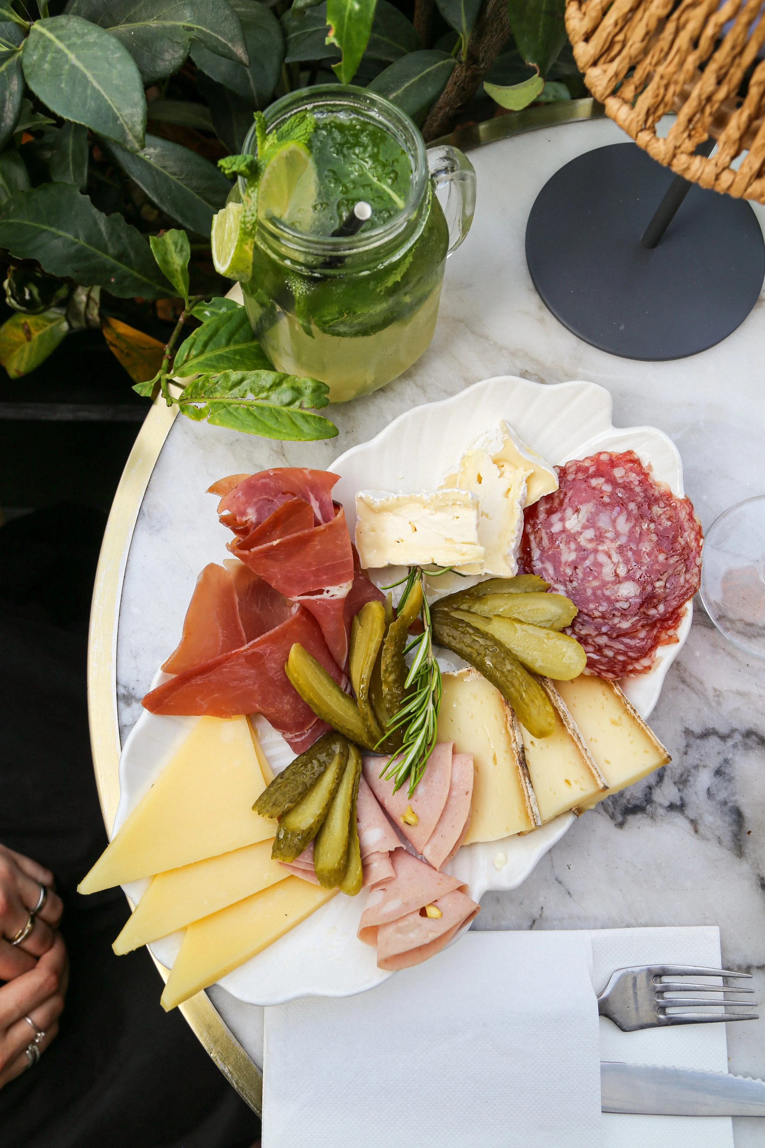 Un plateau de fromages, charcuterie, cornichons, et d'autres accompagnements, avec un verre de cocktail vert à côté, posé sur une table en marbre.