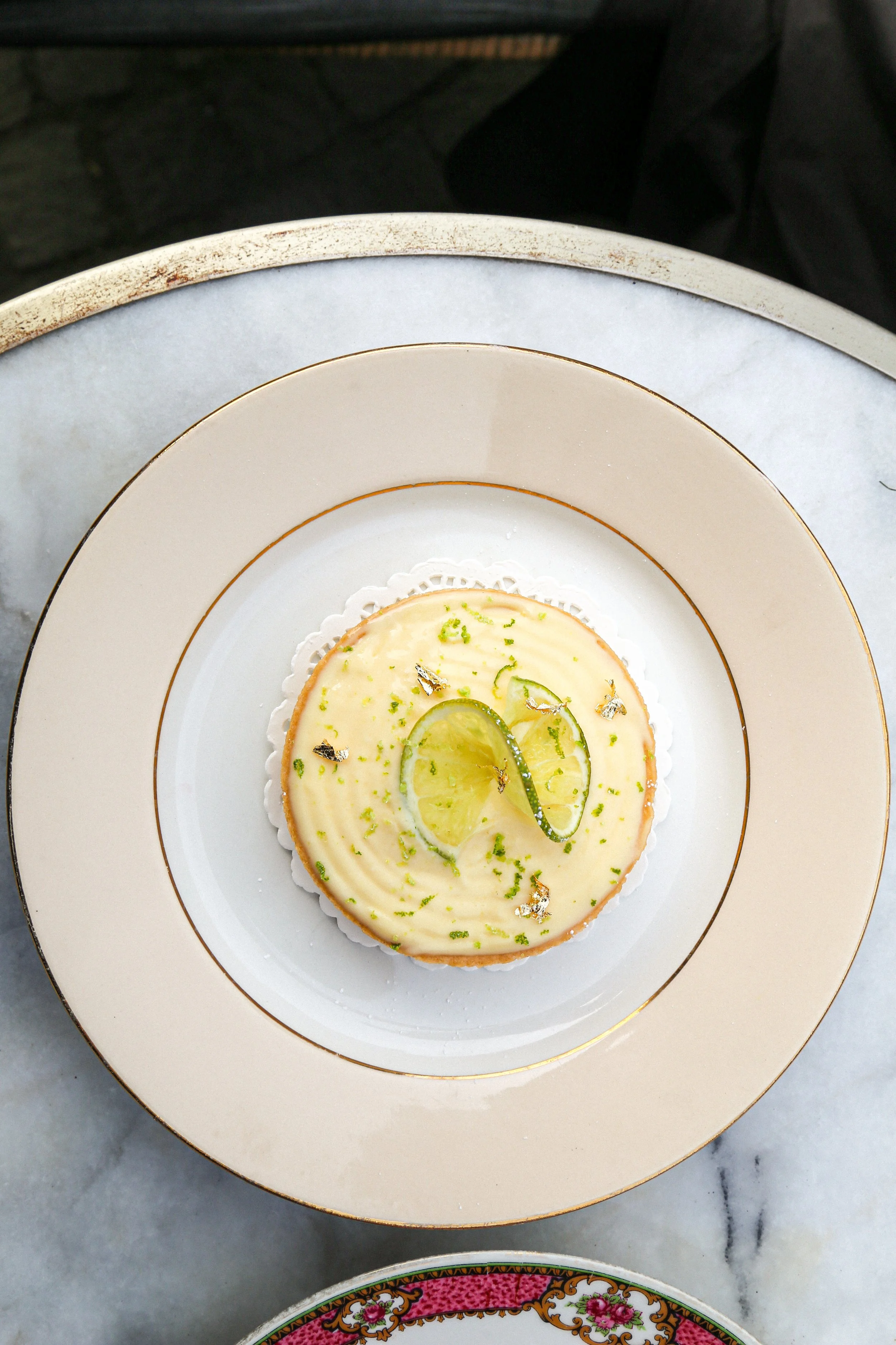 Un dessert crémeux garni de tranches de citron vert et de zestes verts, présenté sur une assiette blanche avec des lignes dorées, posée sur une table en marbre.
