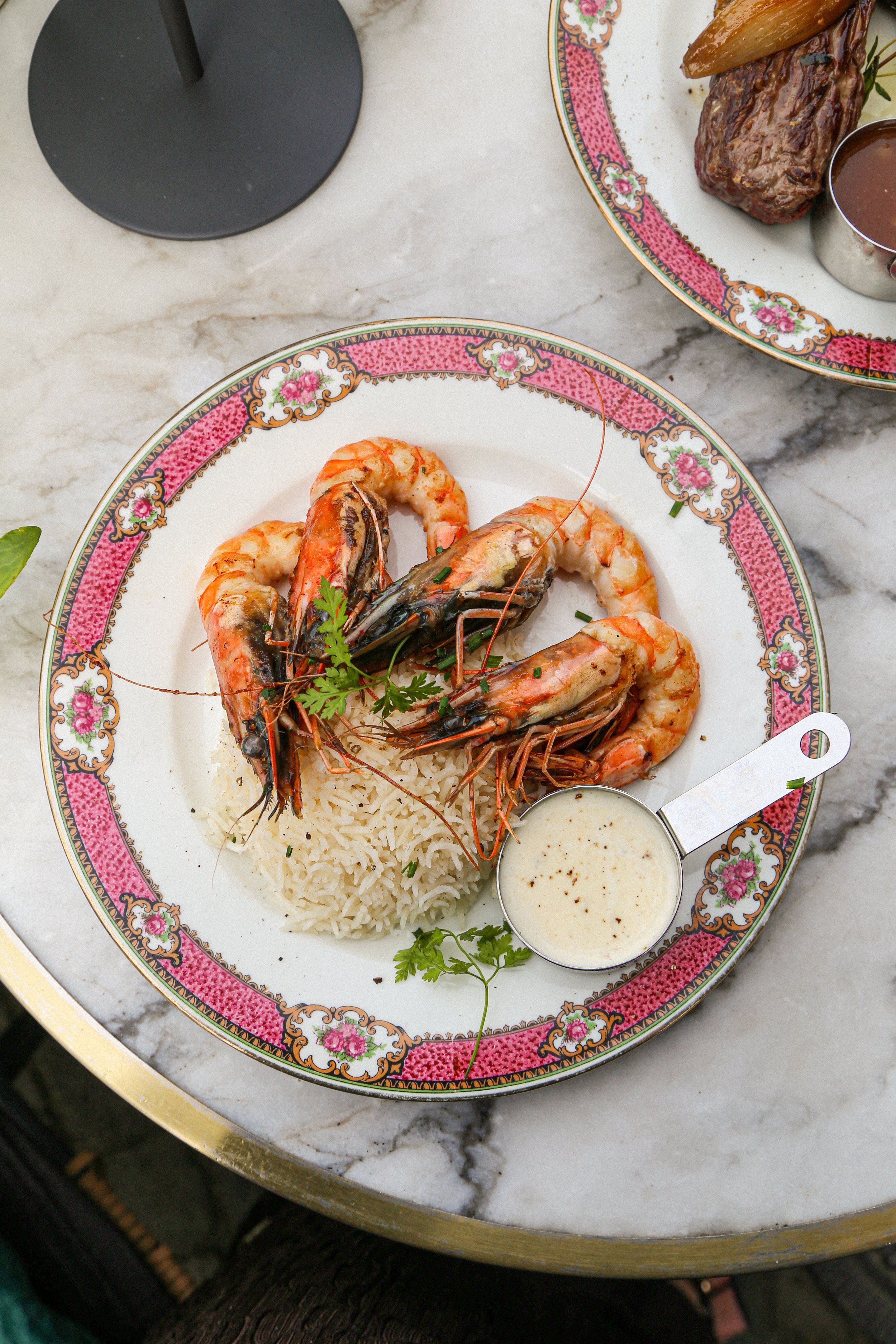 Plate de crevettes avec du riz blanc, accompagnée de sauce, sur une assiette décorée de motifs floraux rose et or, sur une table en marbre.