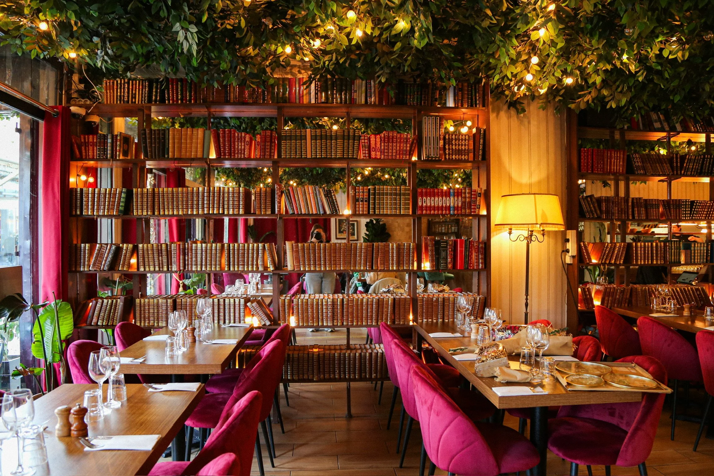 Une salle à manger d'un restaurant avec des tables en bois et des assises en velours rouge, une bibliothèque remplie de livres derrière une paroi en bois, des lampes d'ambiance et des feuilles vertes au plafond, créant une atmosphère chaleureuse et cosy.