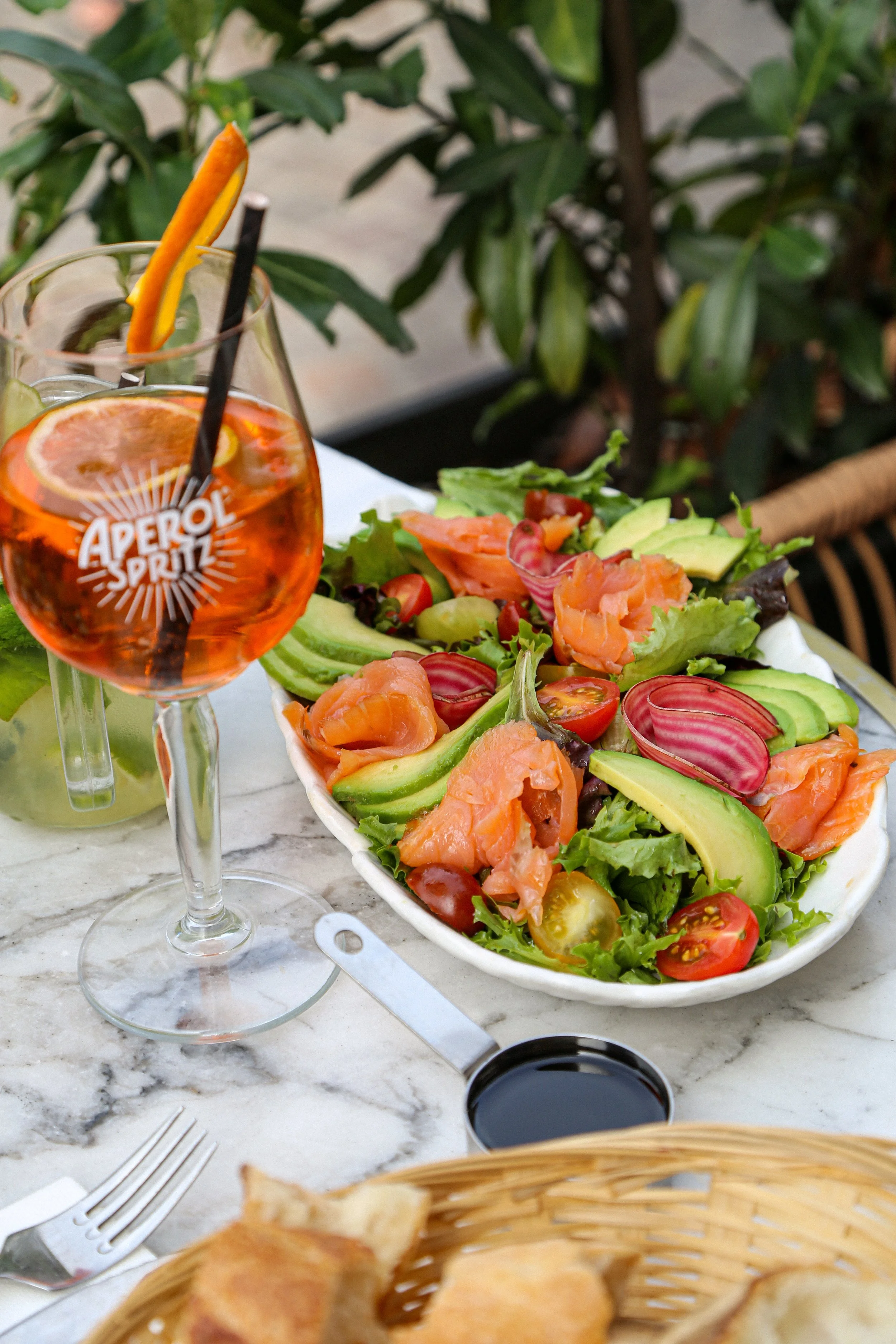 Verre d'Aperol Spritz avec tranche d'orange et paille, assiette de salade comprenant avocat, tomates, saumon, et laitue, au centre une petite tasse de sauce ou vinaigrette, en arrière-plan un fond de plantes vertes, une fourchette et un couteau sont 