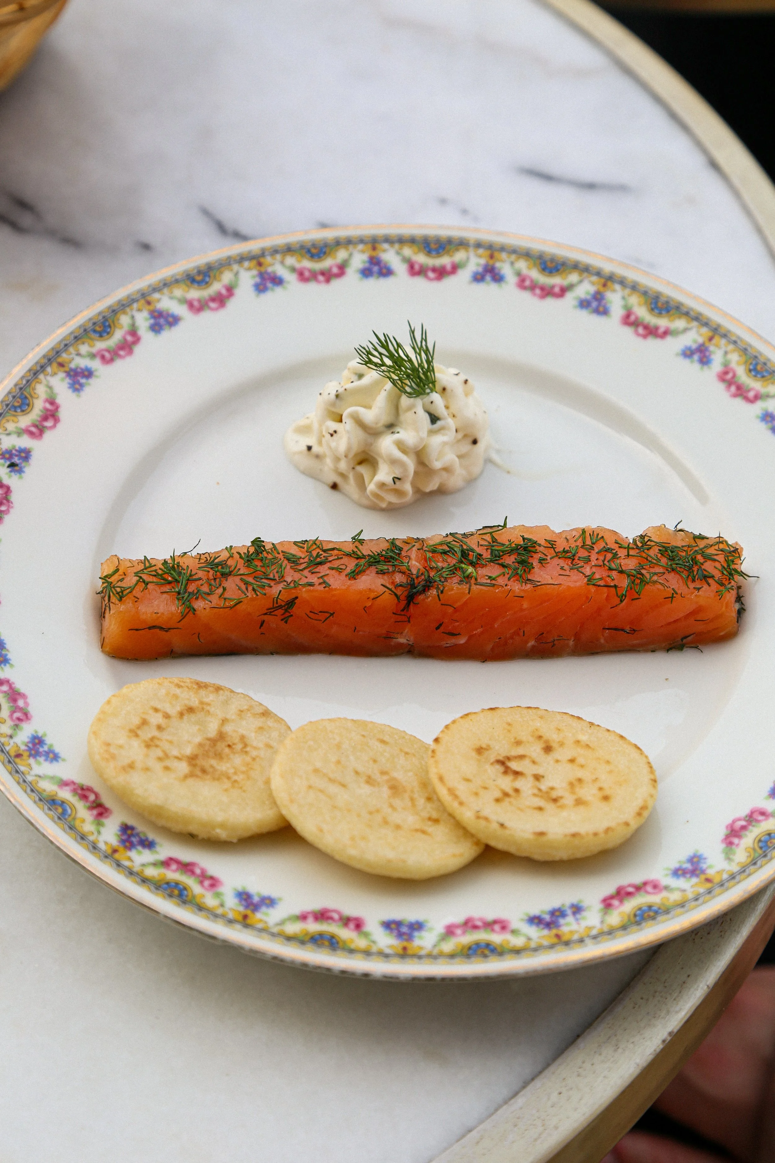 Une assiette en porcelaine blanche avec un motif floral coloré, contenant du poisson fumé garni d'aneth, de pommes de terre petites, rondes et grillées, et une portion de crème avec une branche de dill.