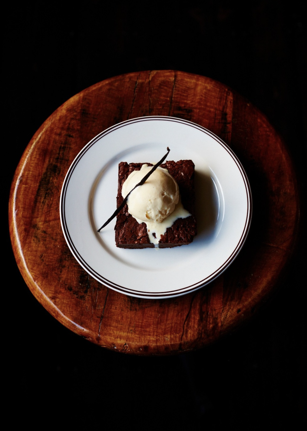 A delicious homemade brownie topped with a scoop of vanilla ice cream on a white plate, garnished with a vanilla bean, on a wooden table.