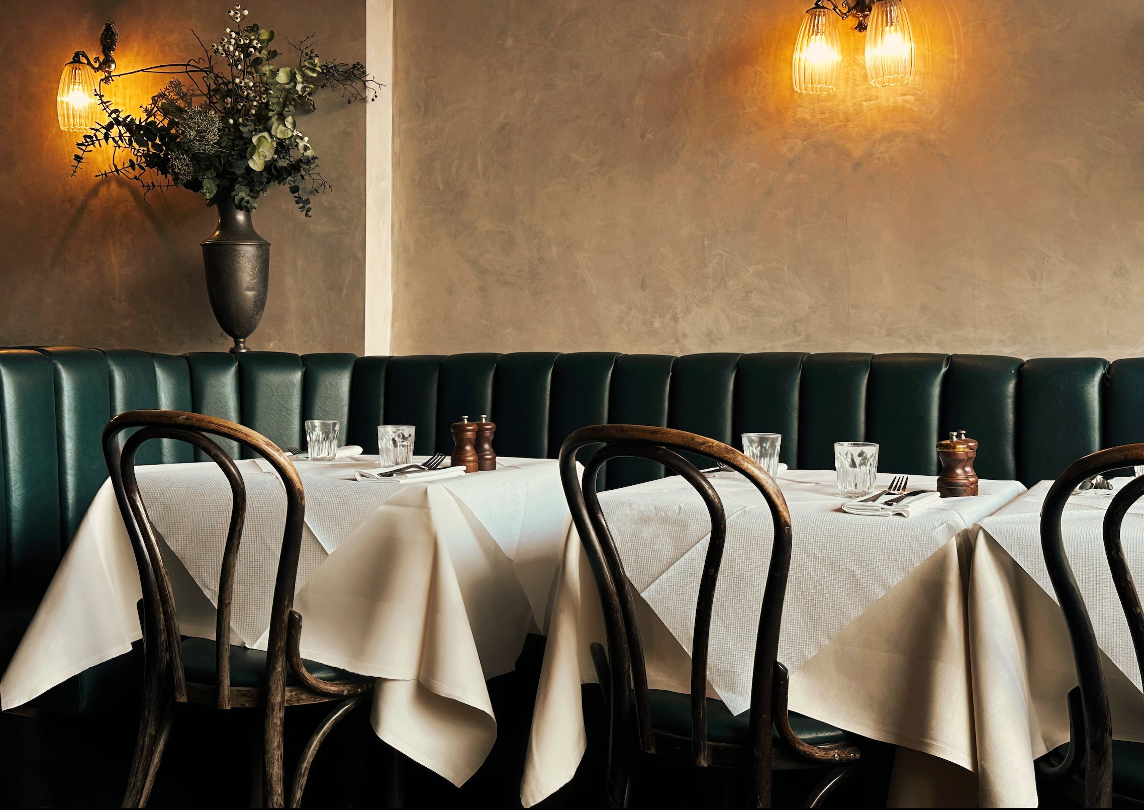 A stunning restaurant interior with tables set with wine glasses, elegant tablecloths, surrounded by wooden bistro chairs and a green leather banquette, with a large beautiful floral arrangement and warm wall lighting.