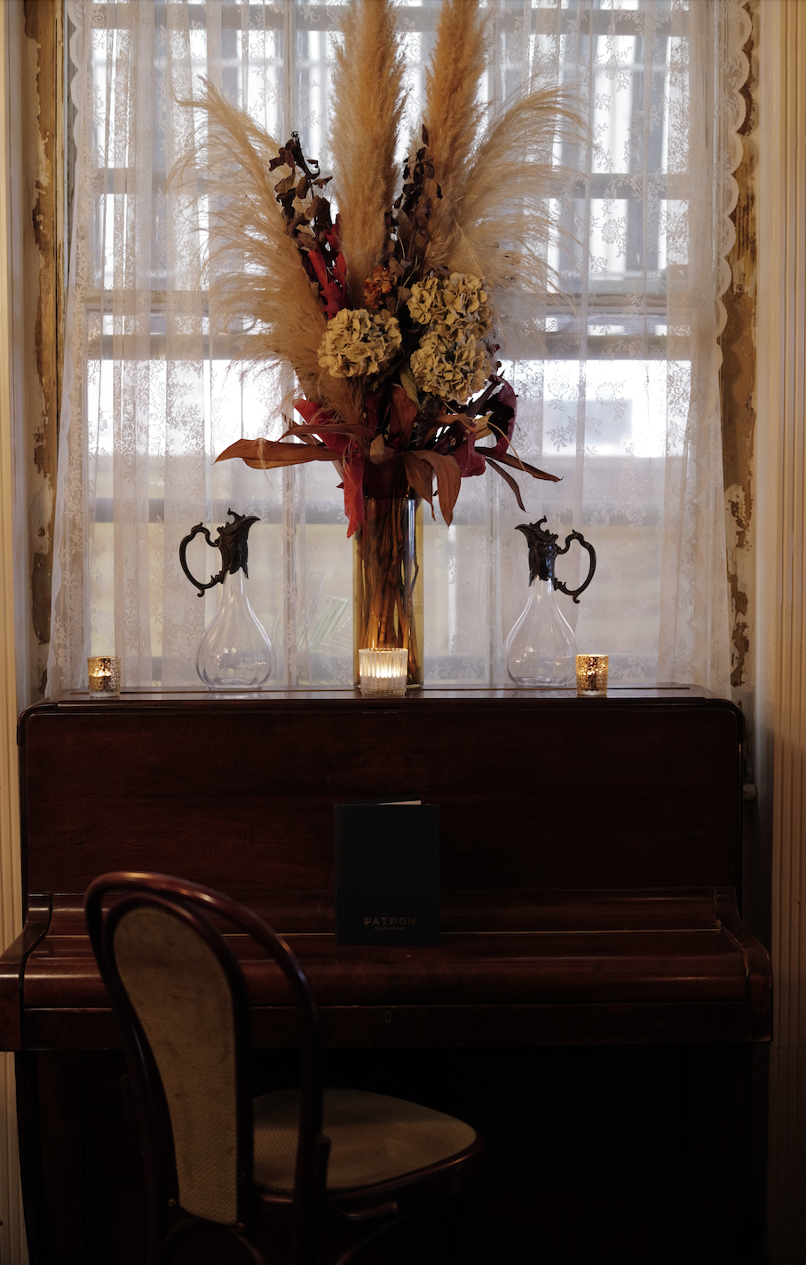 A stunning vintage piano set against a window with lace curtains, with a large floral arrangement and three small candle holders with lit candles at Patron's 1920s Spirit Lounge.