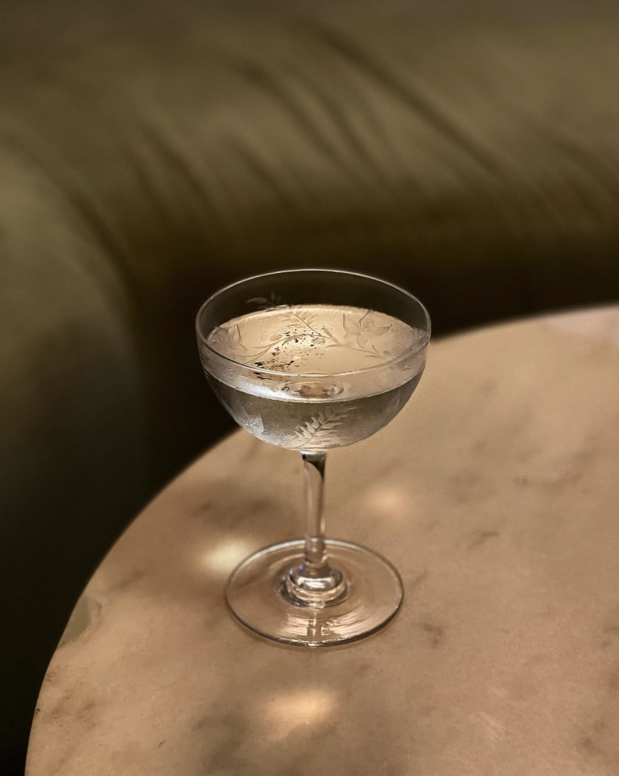 A signature Dirty Martini cocktail, served in a vintage coupe glass, placed on a marble table surrounded with plush velvet seating.