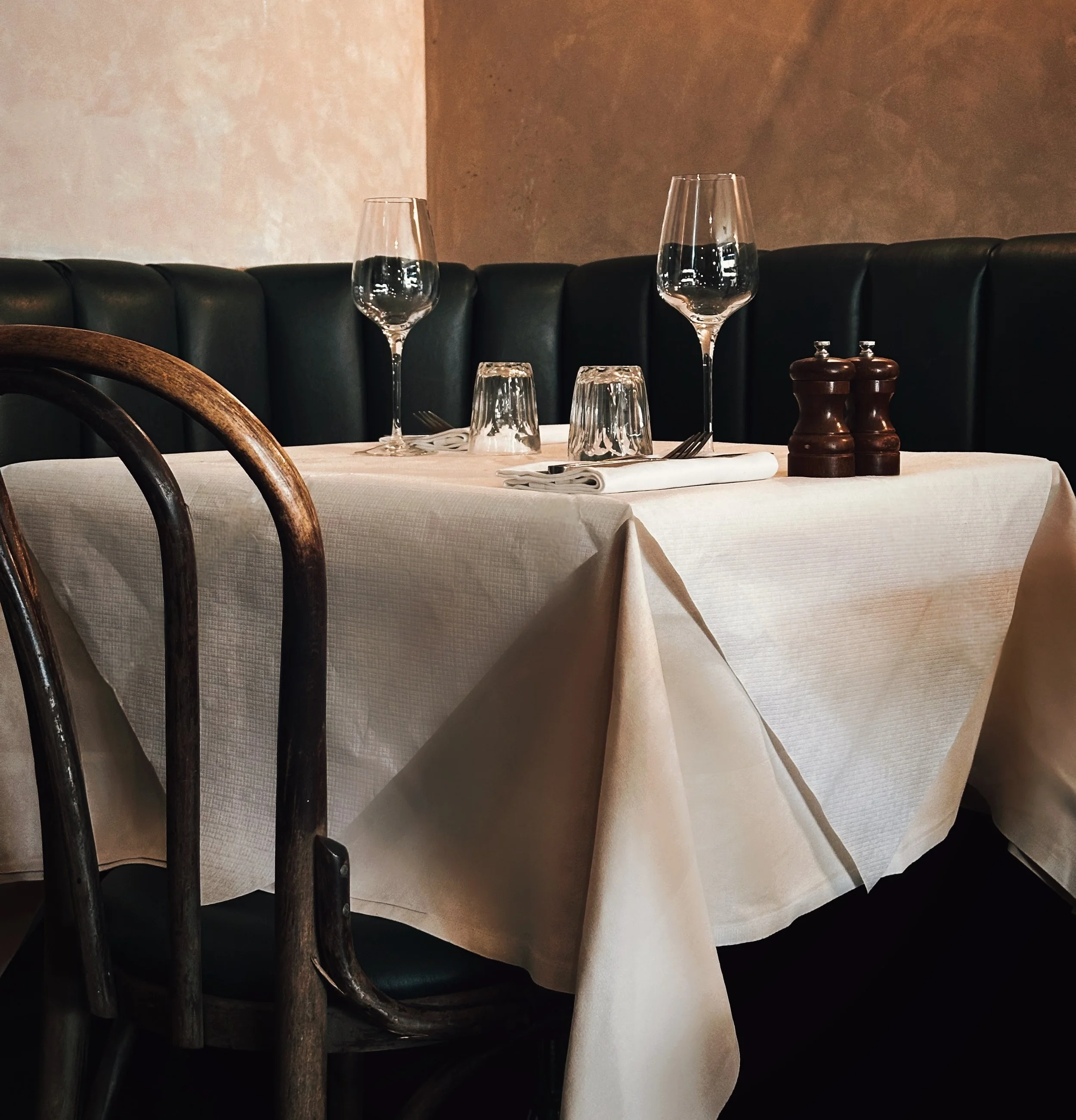 Beautifully set dinner table with elegant tablecloth, wine glasses and neatly folded napkins, at the bar room at Patron restaurant, with a dark green leather banquettes and classic bistro chairs.