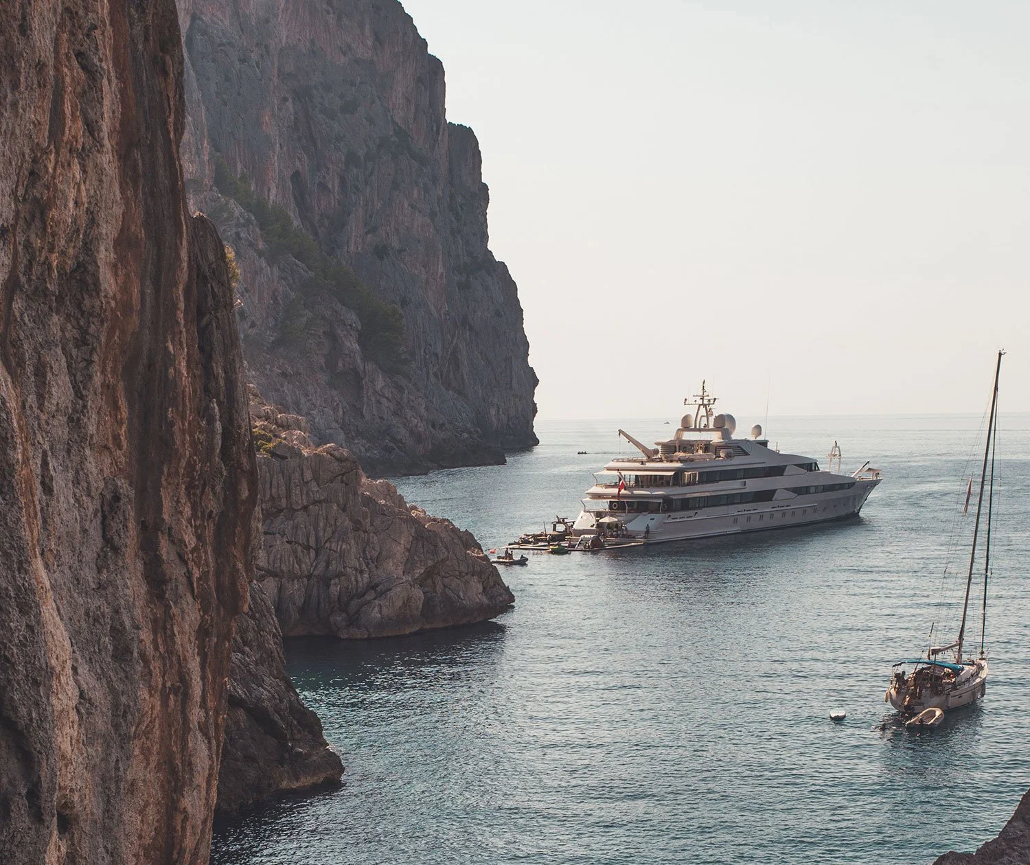 Private yacht charter: Bahamas, Caribbean, Mediterranean, Southeast Asia. Luxury yacht at anchor beneath steep coastal cliffs.