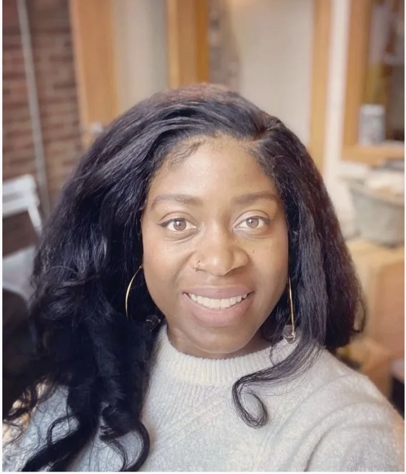 Close-up of a smiling woman with long dark wavy hair wearing hoop earrings and a light-colored sweater, indoors with a brick wall and wooden cabinets in the background.