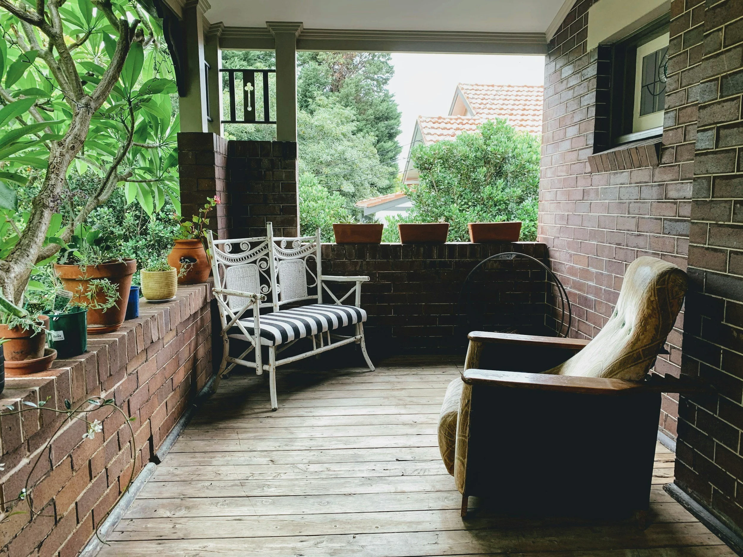 Covered porch or patio area with potted plants, outdoor chairs including a white metal chair with striped cushion, and wooden chairs, surrounded by brick walls and greenery outside.