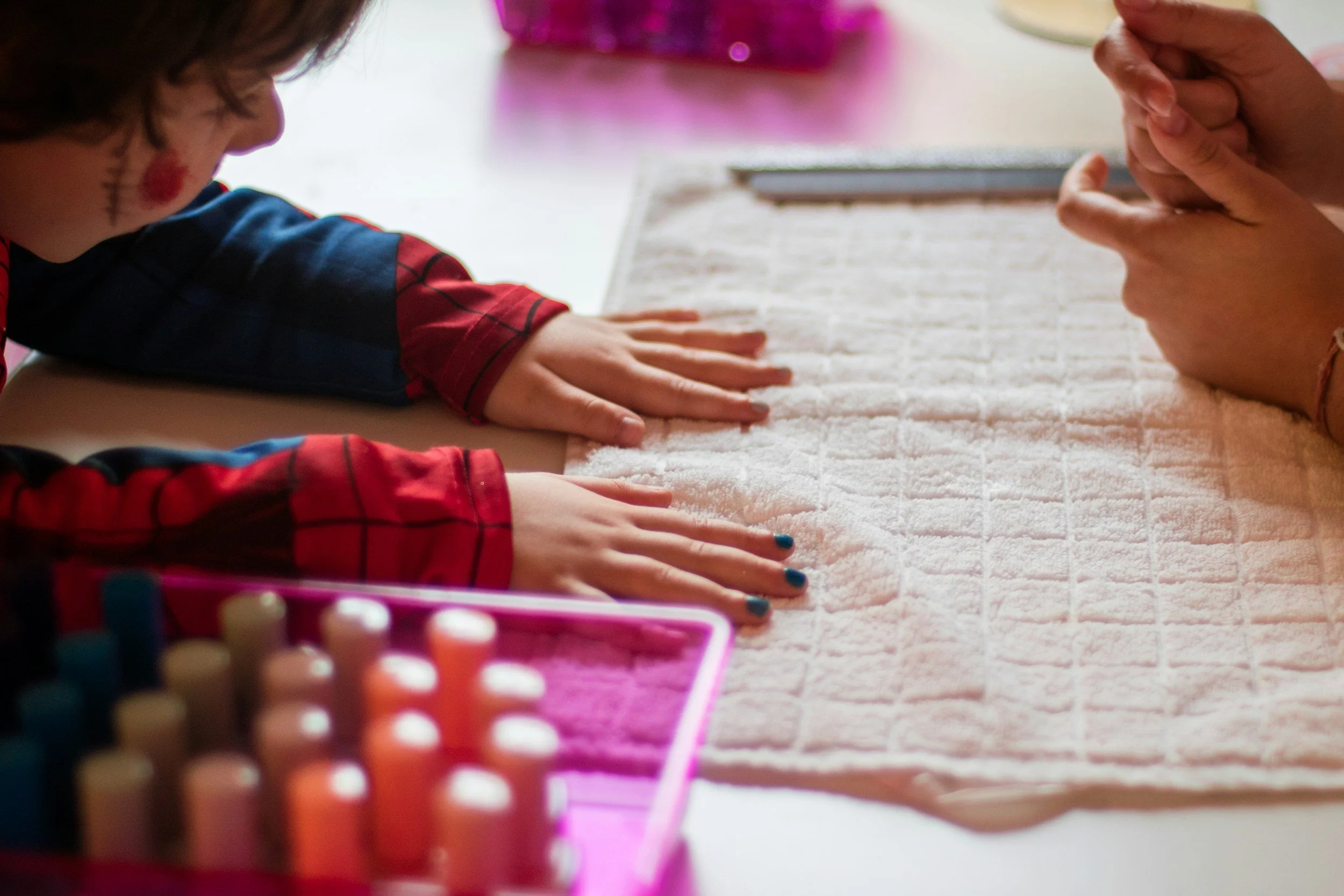 Child and adult with hands pressed on a white textured surface, seen from the side, with colorful art supplies in the foreground.