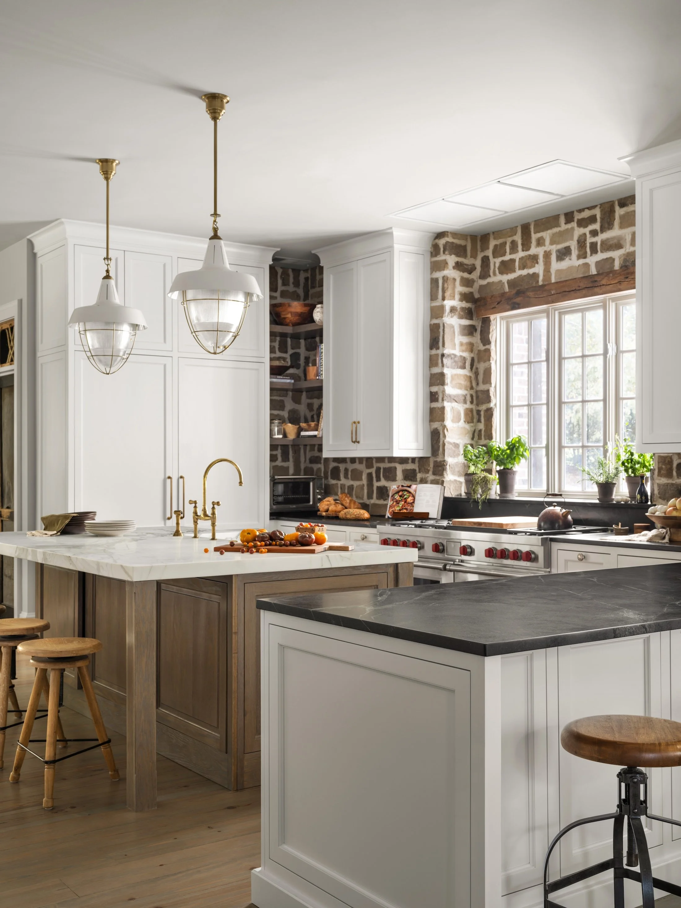 Bright custom kitchen with white cabinetry, wood island, stone walls, pendant lighting, and large windows.